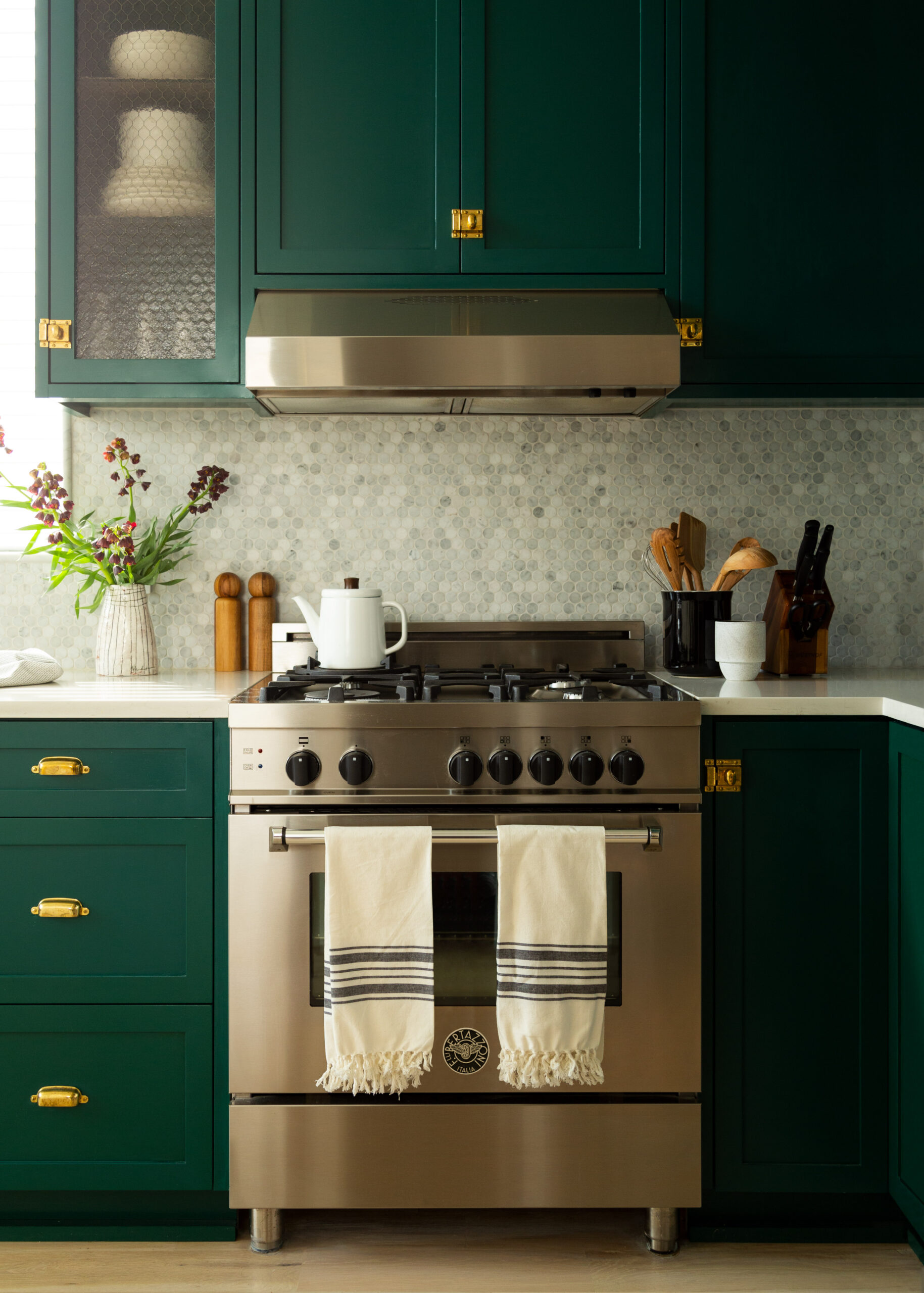 Deep green cabinets with brass hardware and a stainless range create a rich, modern kitchen accented by a soft mosaic backsplash.