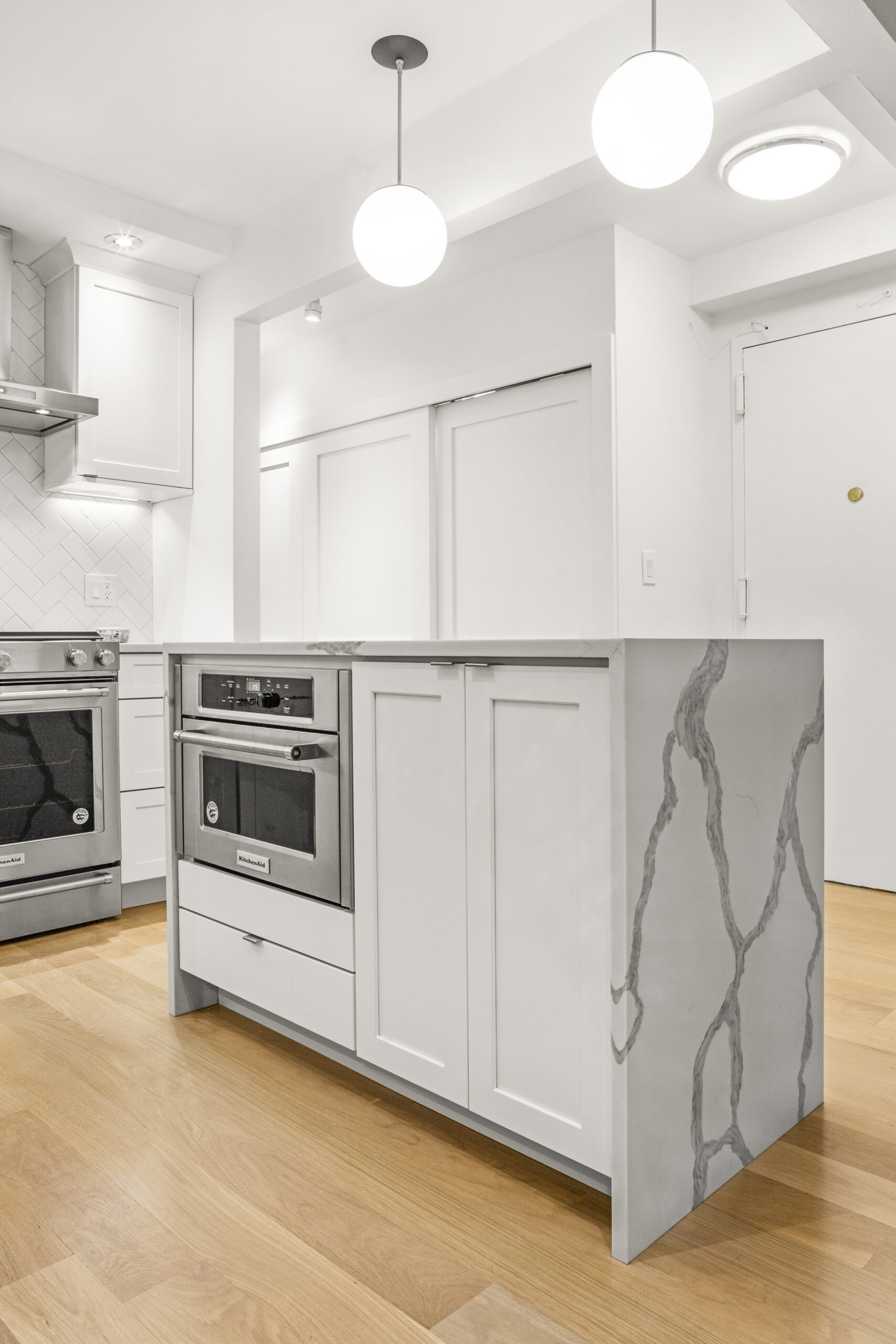 Compact white island with built in oven, marble waterfall sides, globe pendants above, and light wood flooring beside a matching range wall.