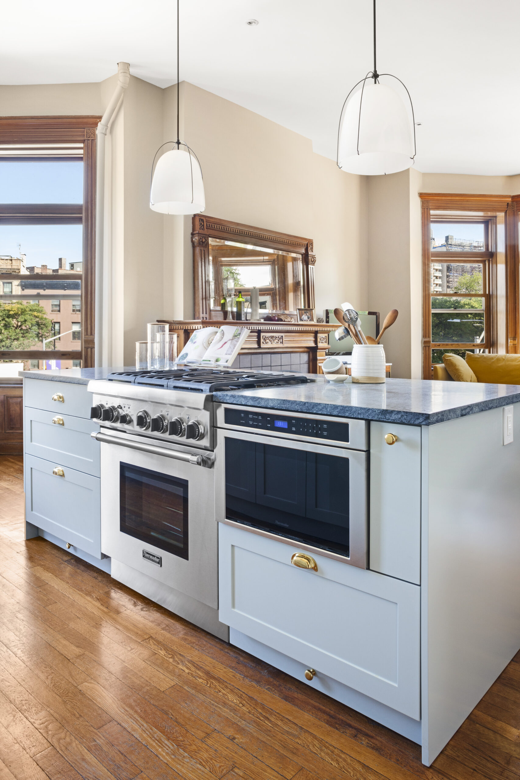 Blue island with range and microwave, brass pulls, wood flooring, and large windows looking toward a traditional living area with carved mirror.