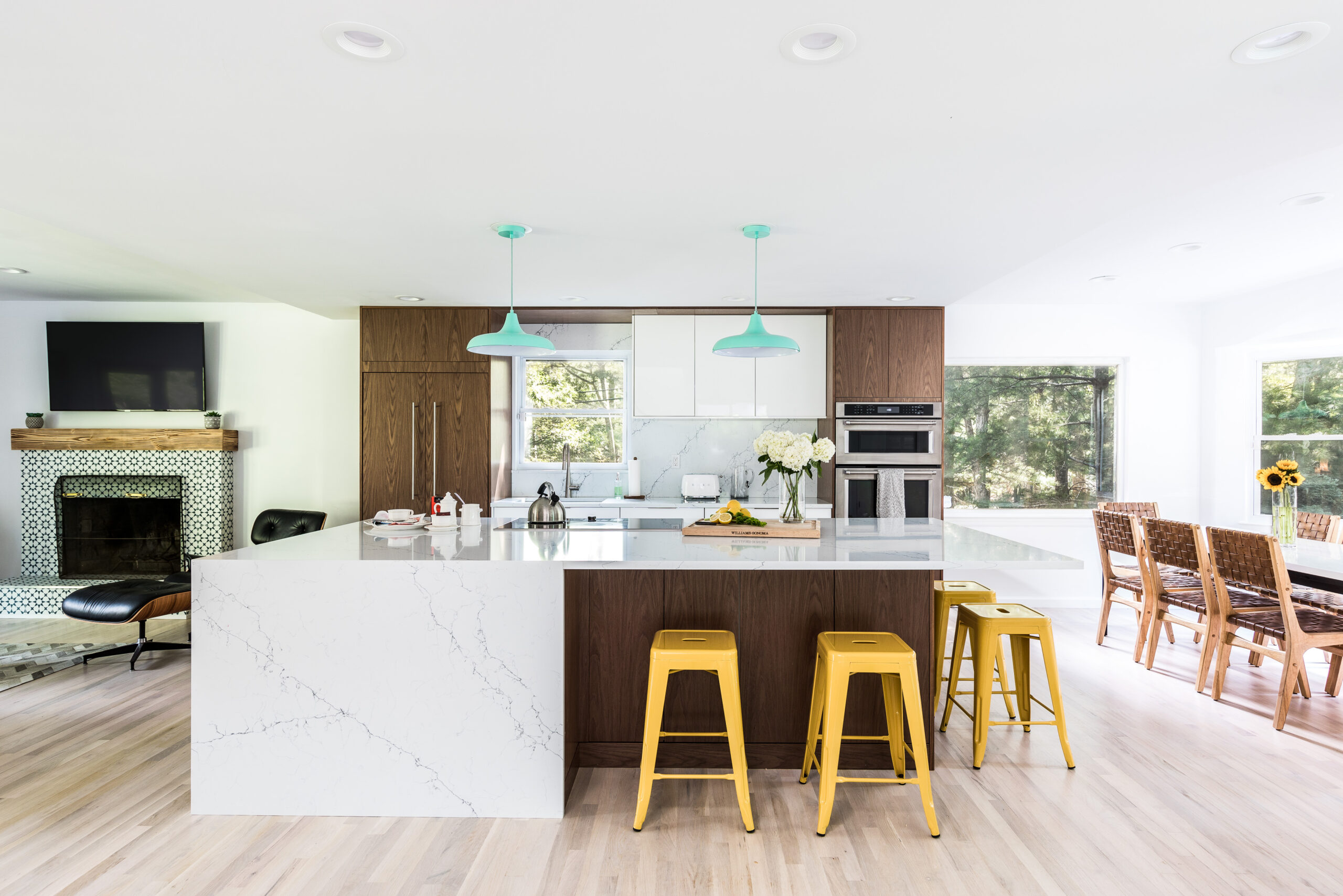 Open concept kitchen with white and walnut cabinets, a large waterfall island, yellow metal stools, aqua pendants, and views toward fireplace and dining.