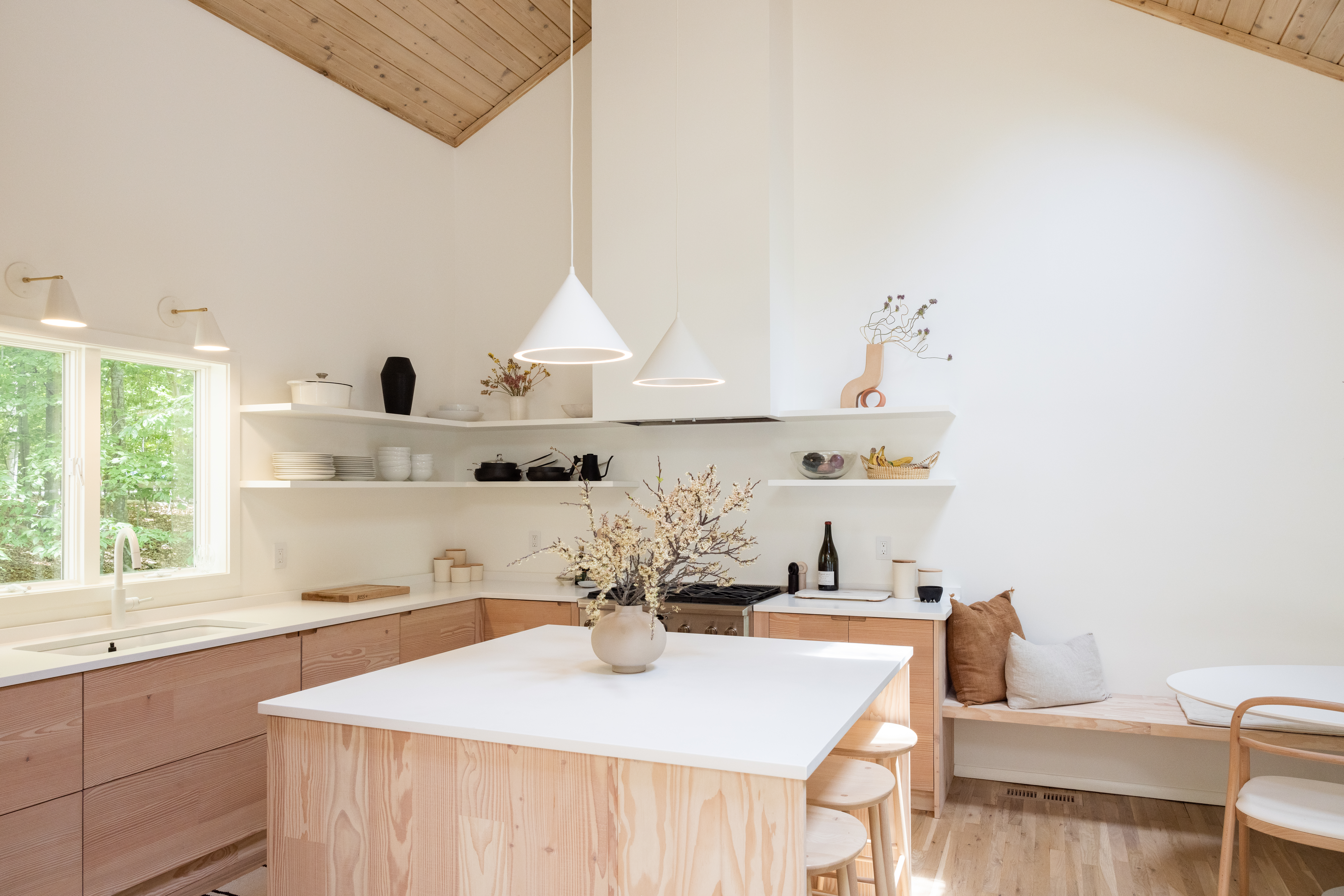 Light Scandinavian style kitchen with pale wood cabinets, matching island with white top, open shelves, and simple stools around a built in bench.