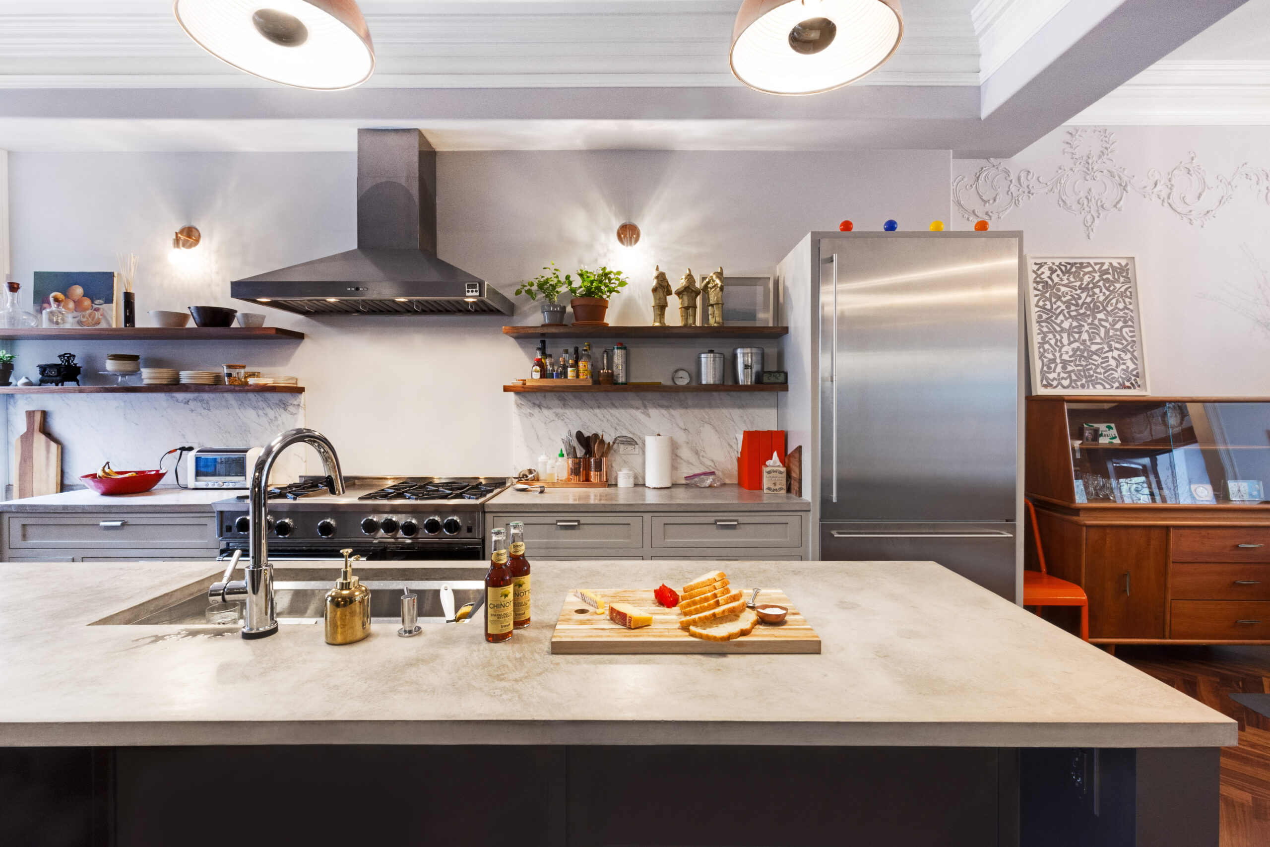 Concrete look island with prep sink in foreground, open shelves and range wall behind, and bread, cheese, and sauces styled on the countertop.