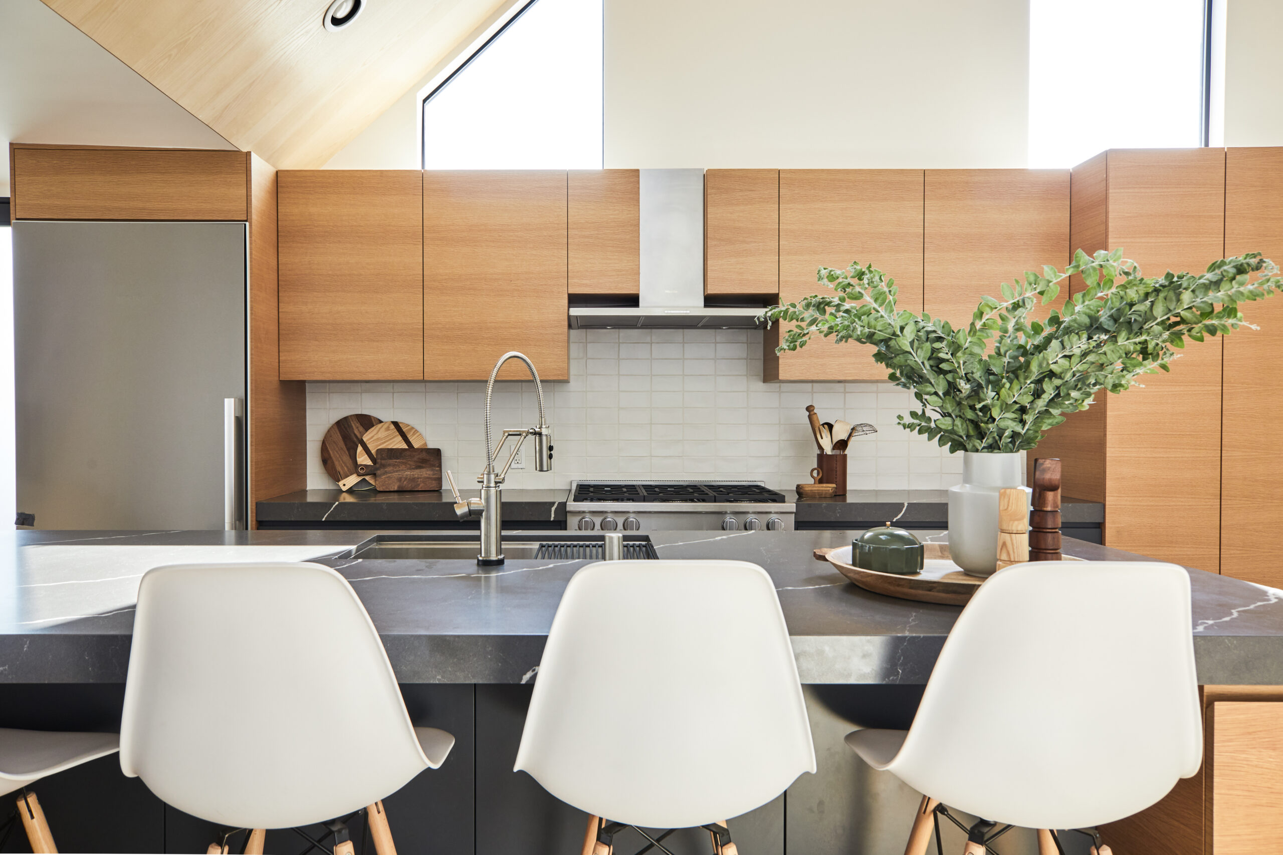 Modern space with dark stone kitchen island, three white shell stools, flat panel oak cabinets, and high windows bringing light onto the work area.