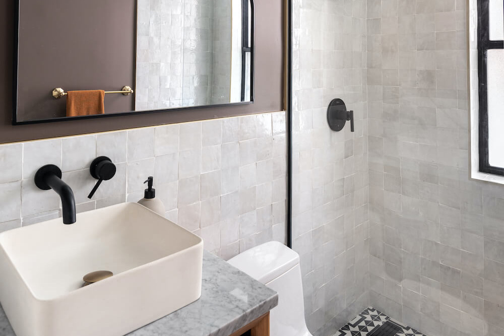 Glossy zellige tile wraps Chelsea’s compact bath, bouncing light around the shower and vanity as matte black fixtures ground the design each day