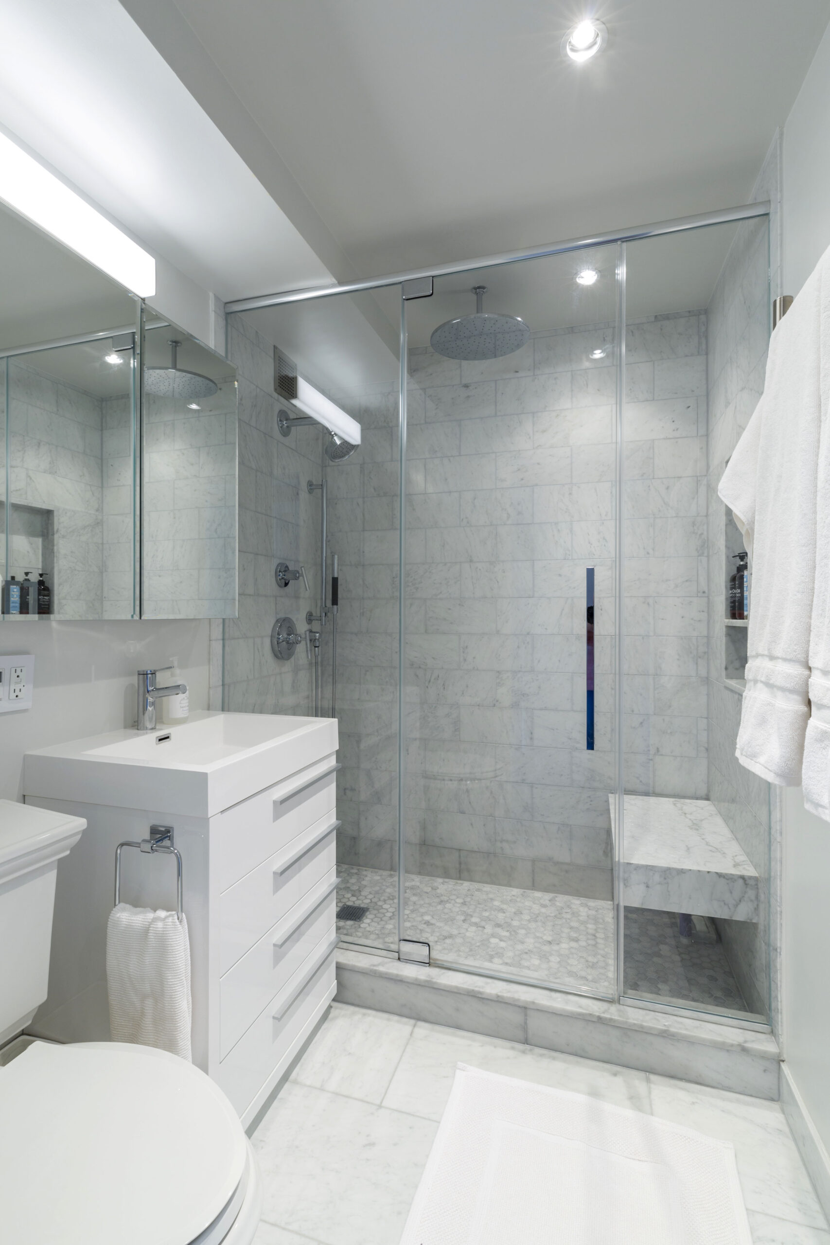 Marble walk-in shower with glass doors, rain head, bench, and white vanity in a bright contemporary bathroom.