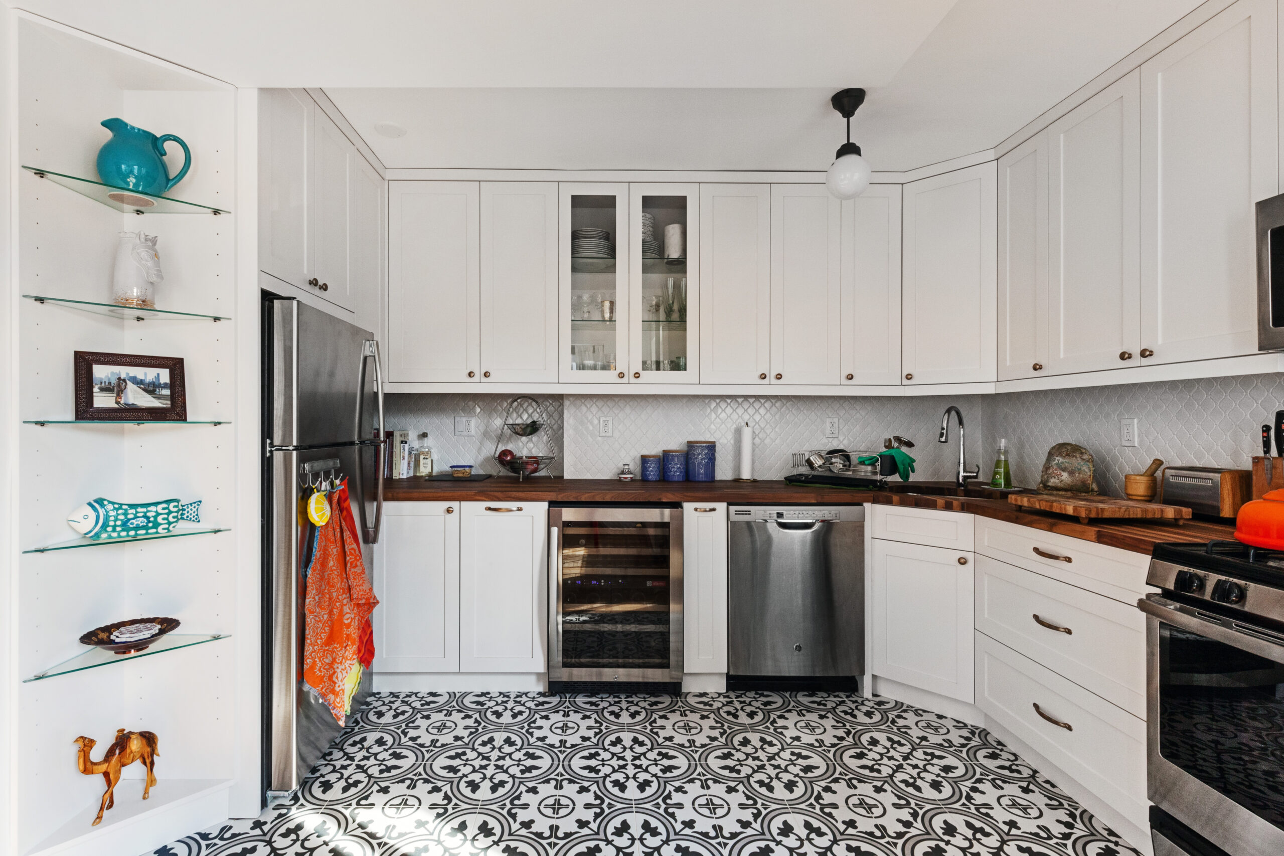 White L shaped kitchen with glass front cabinets, patterned black and white floor and rich butcher block countertops surrounding the appliances.