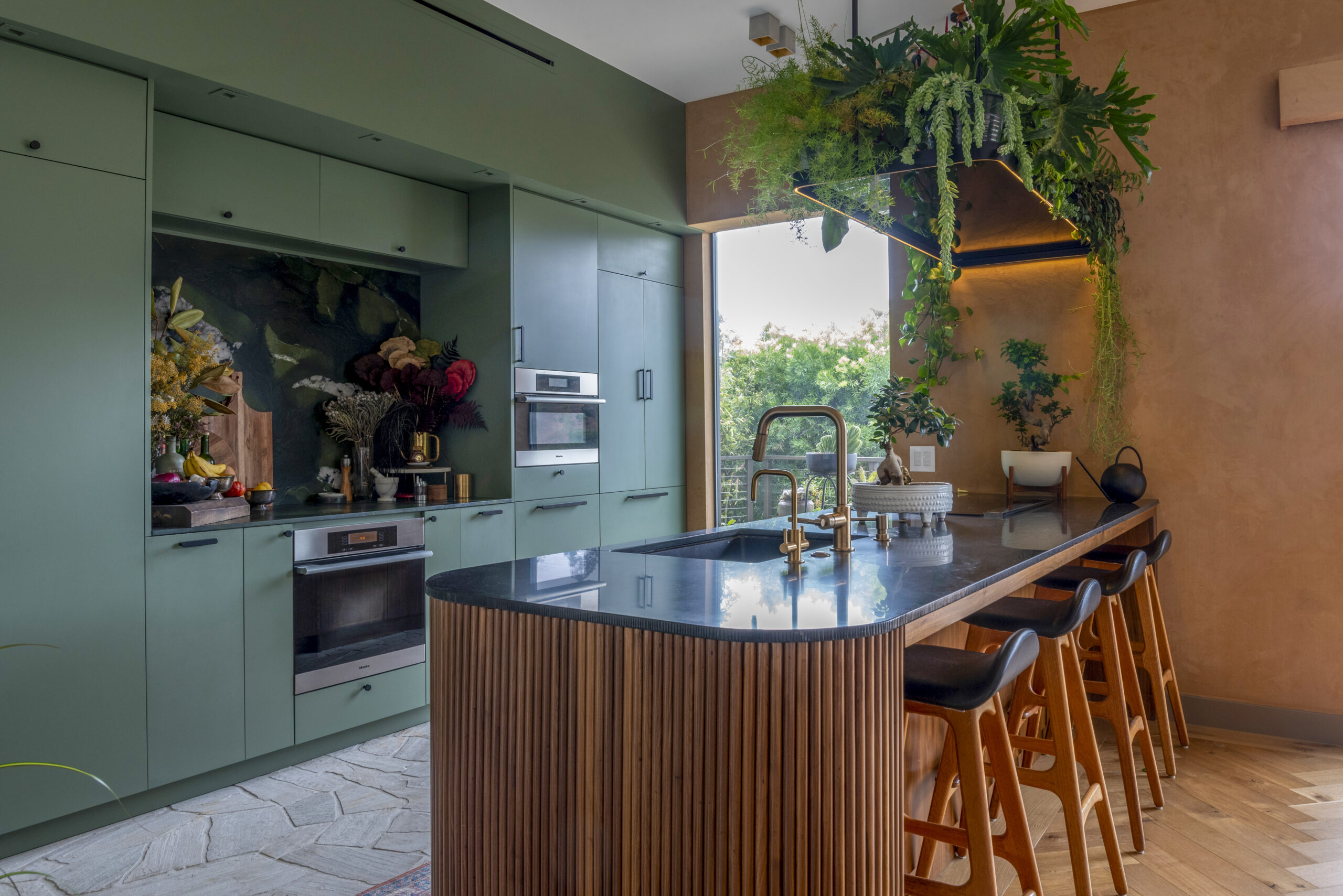 Dramatic green cabinet kitchen with ribbed wood island, dark stone counter, brass fixtures, and hanging plants forming a lush canopy over seating.
