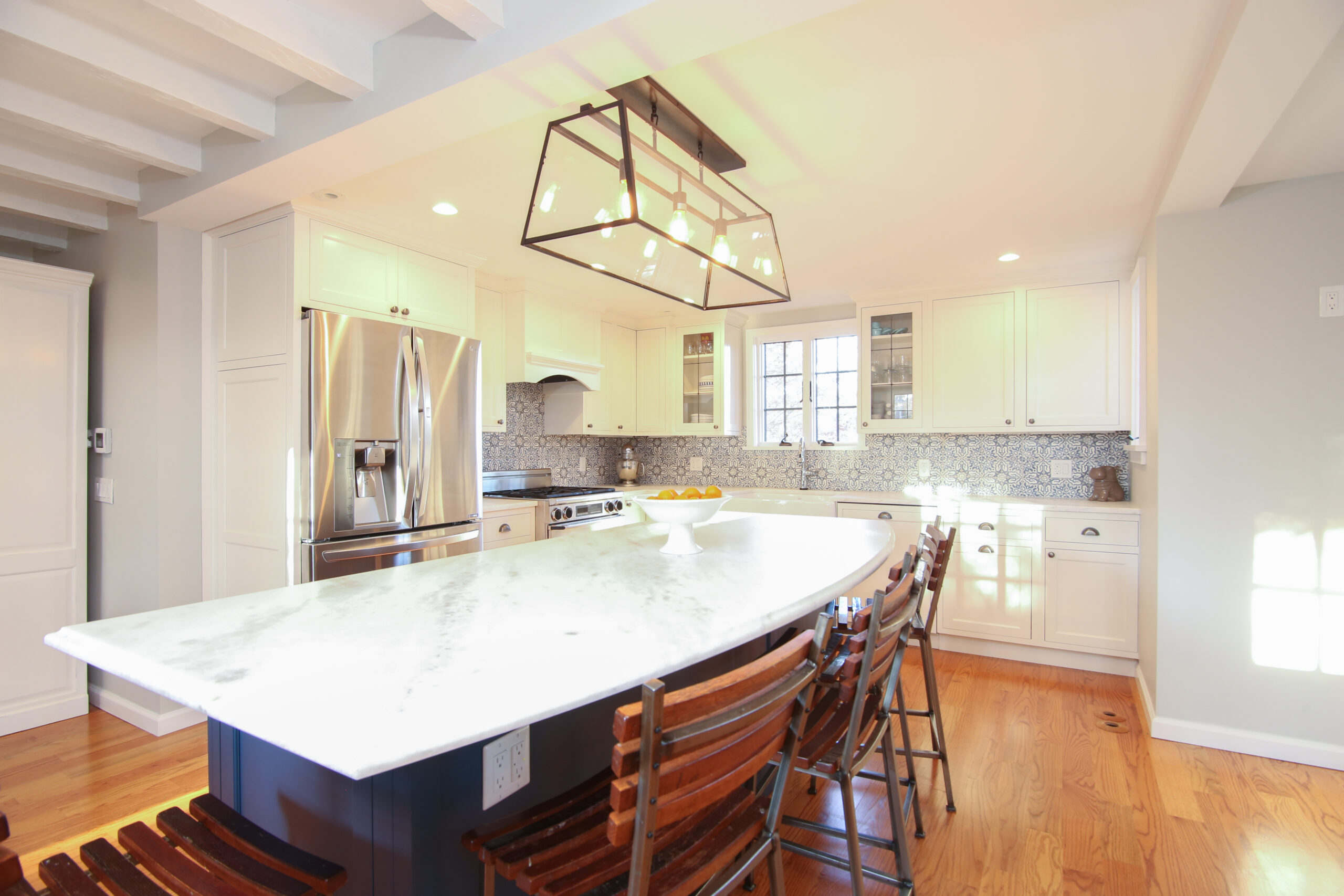 Bright kitchen with curved marble island, dark base and wood stools, white cabinets, stainless fridge, blue patterned backsplash, and glass light.