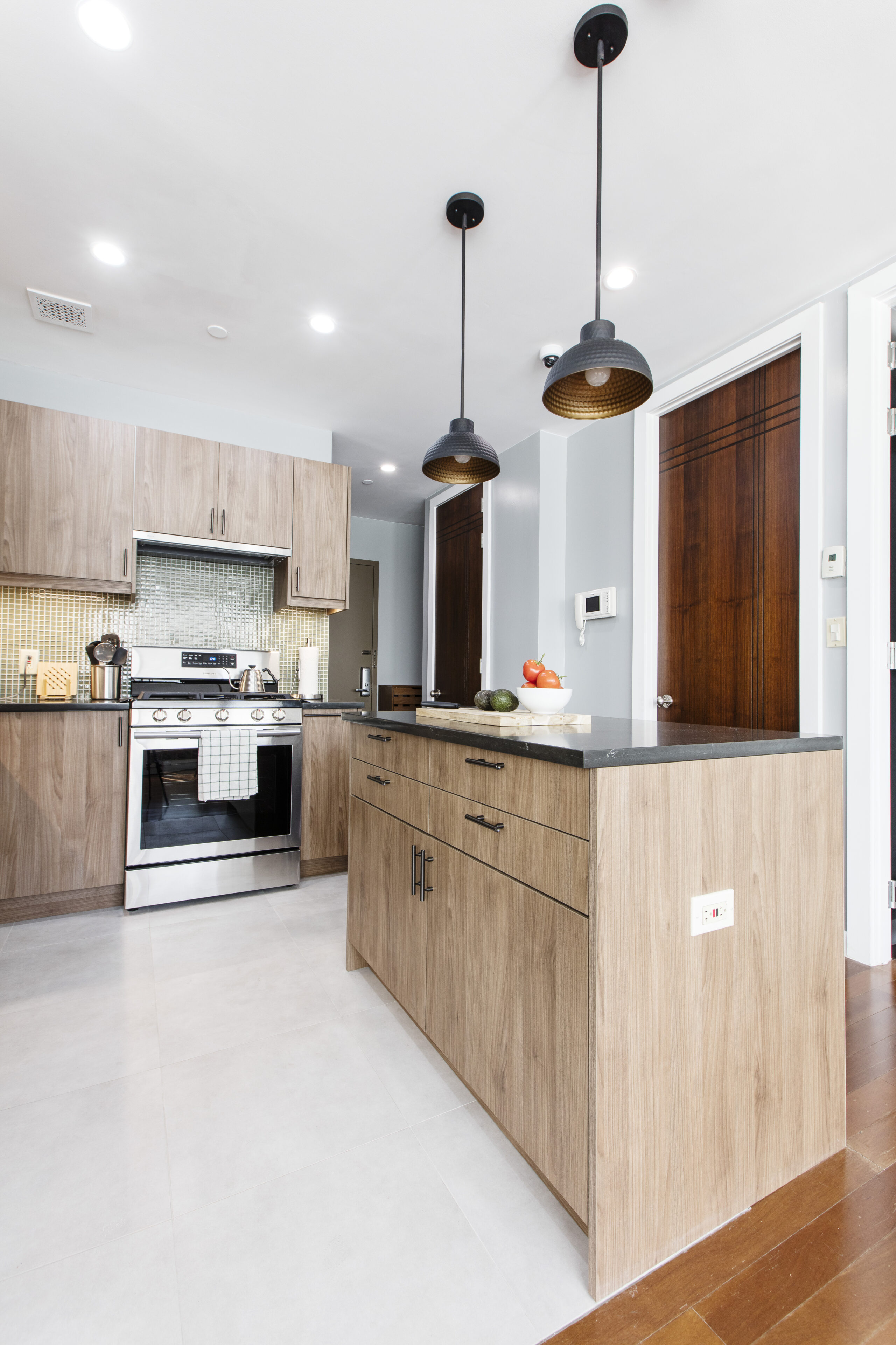 Modern kitchen with flat-front wood cabinets, matching island with dark countertop, and black dome pendants over a bright tile floor.