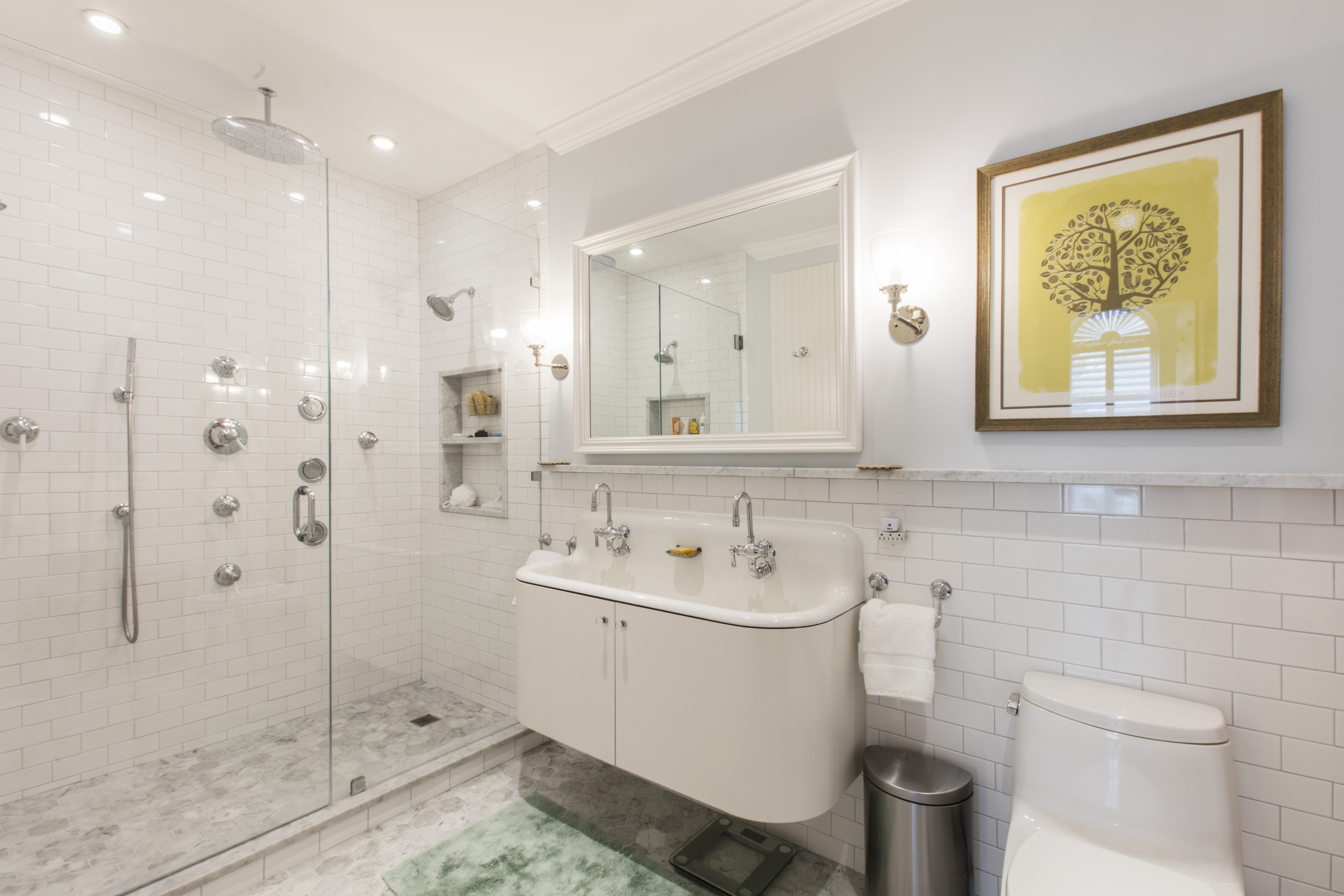 Spacious white subway tile shower with body sprays and rain head beside a double console sink and sconces in a bright bath.