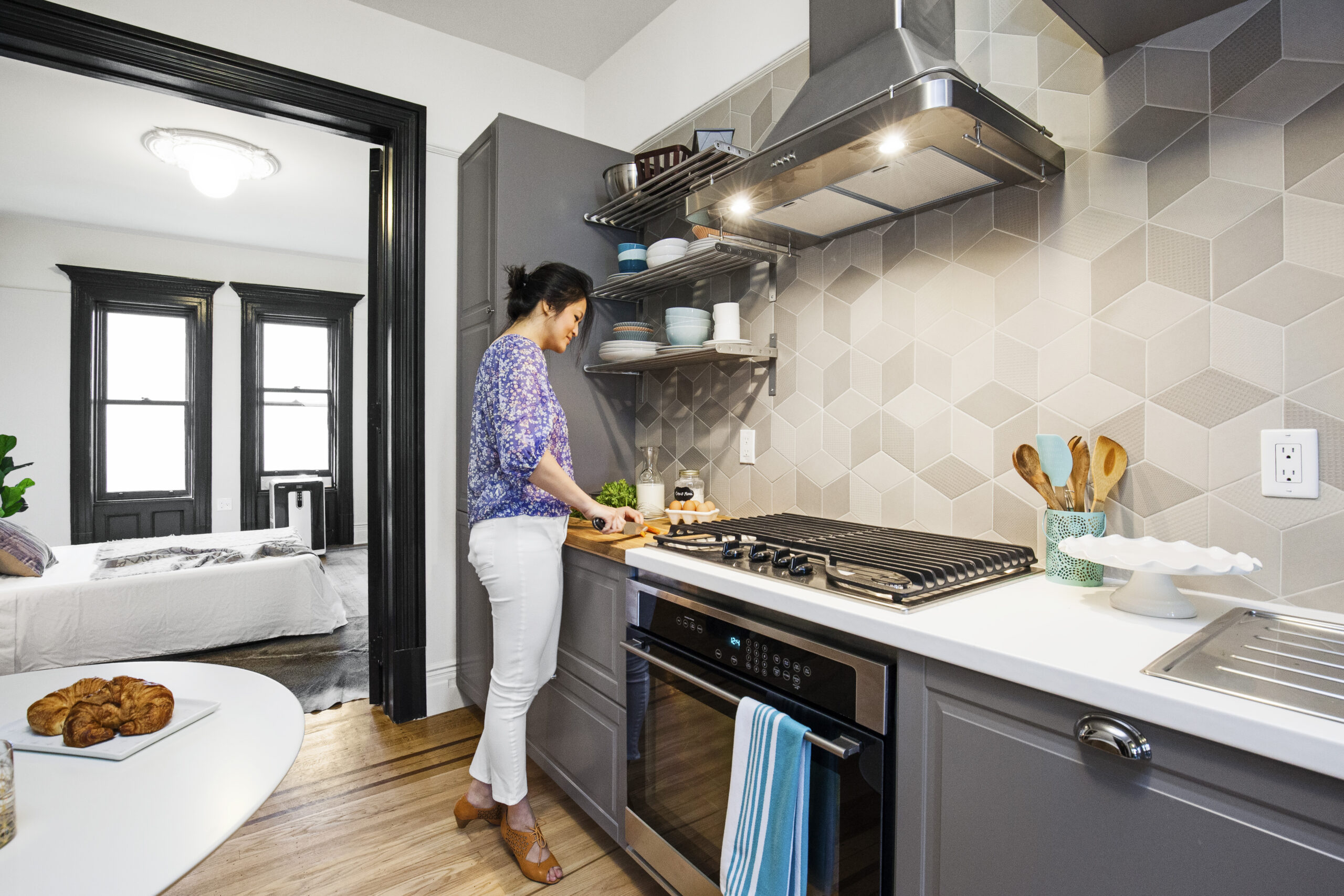 Compact gray kitchen with a cook standing at the range, chopping on a small butcher block cutting board beside a geometric tile backsplash.