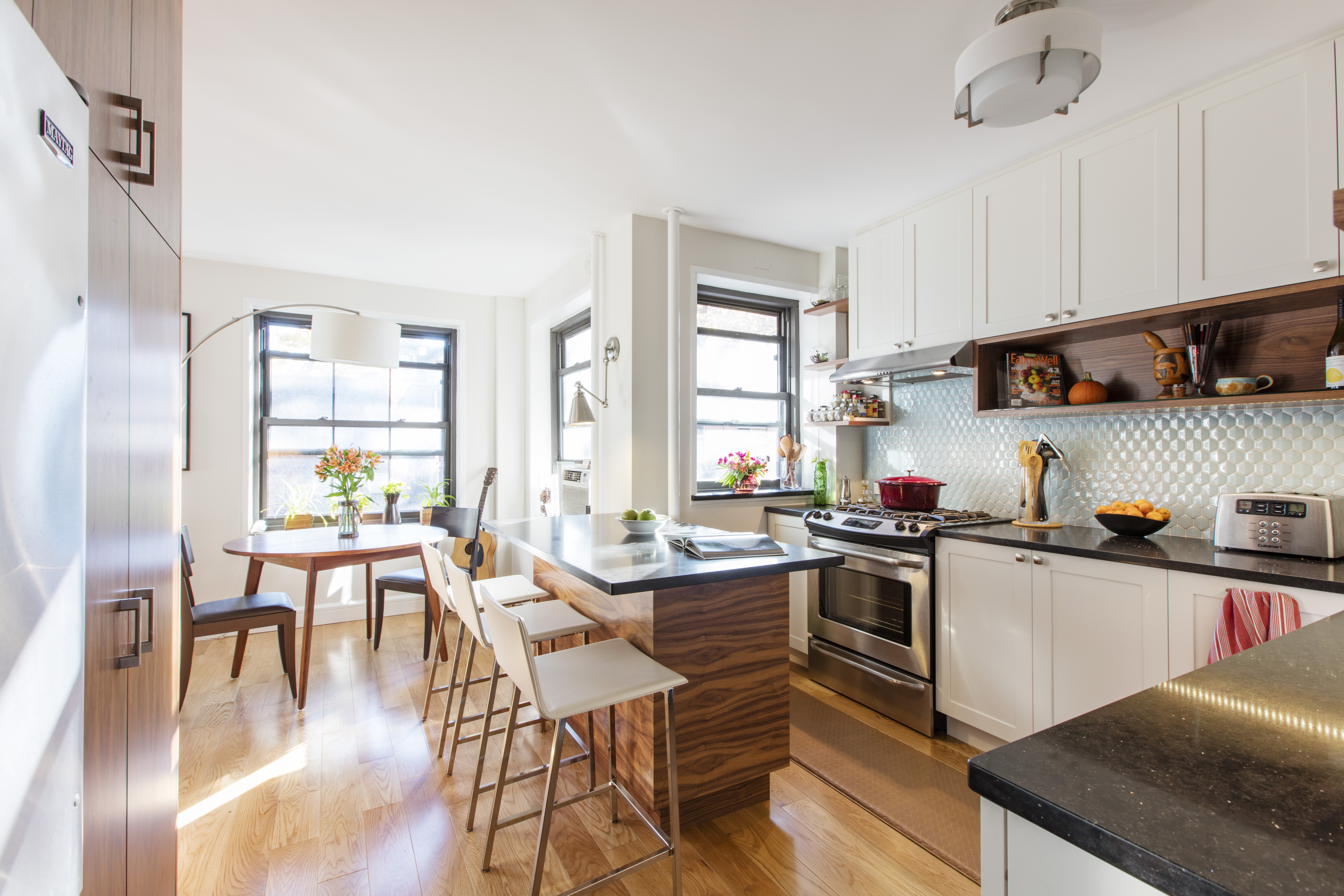 Sunny apartment kitchen with wood-grain kitchen island and stools, white cabinets, open shelves, and a small dining table by the windows.