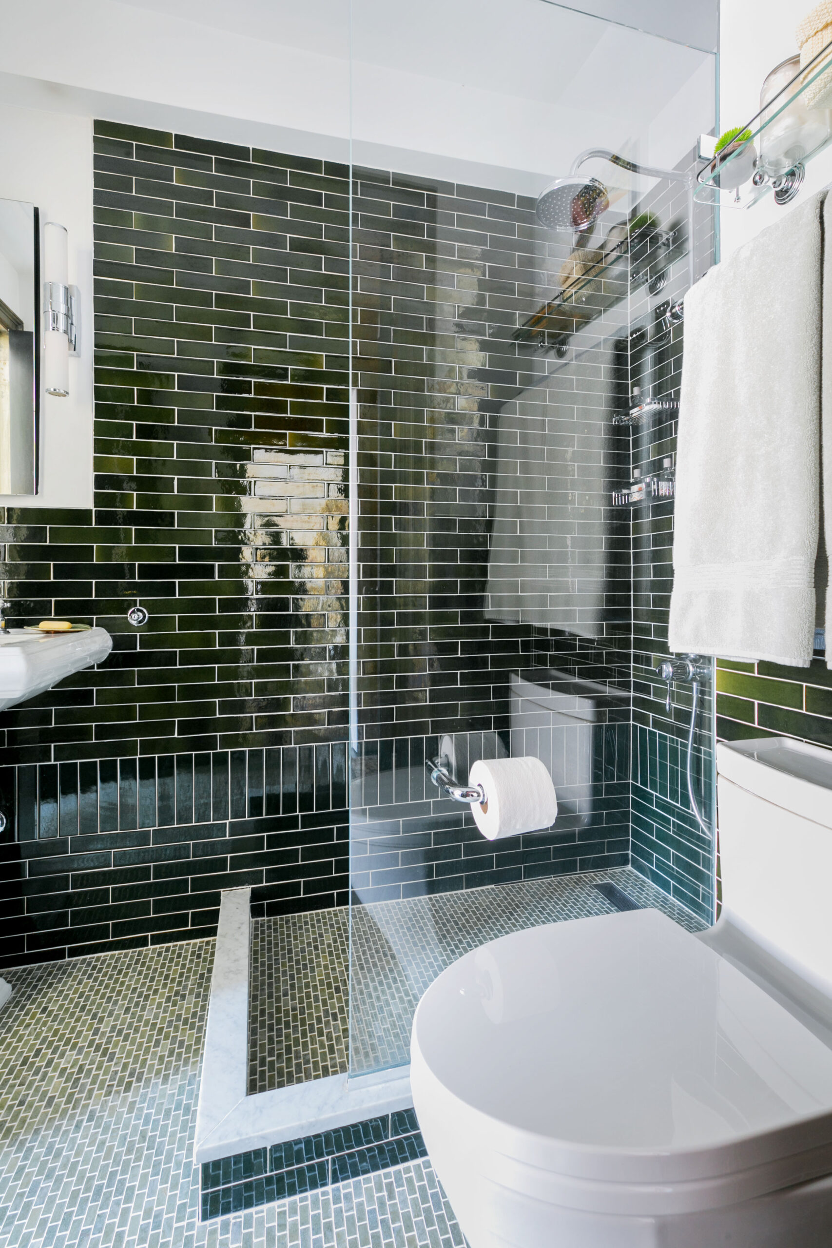 Small bathroom with glossy green subway tile walls, glass shower with marble curb, and mosaic floor tiles.