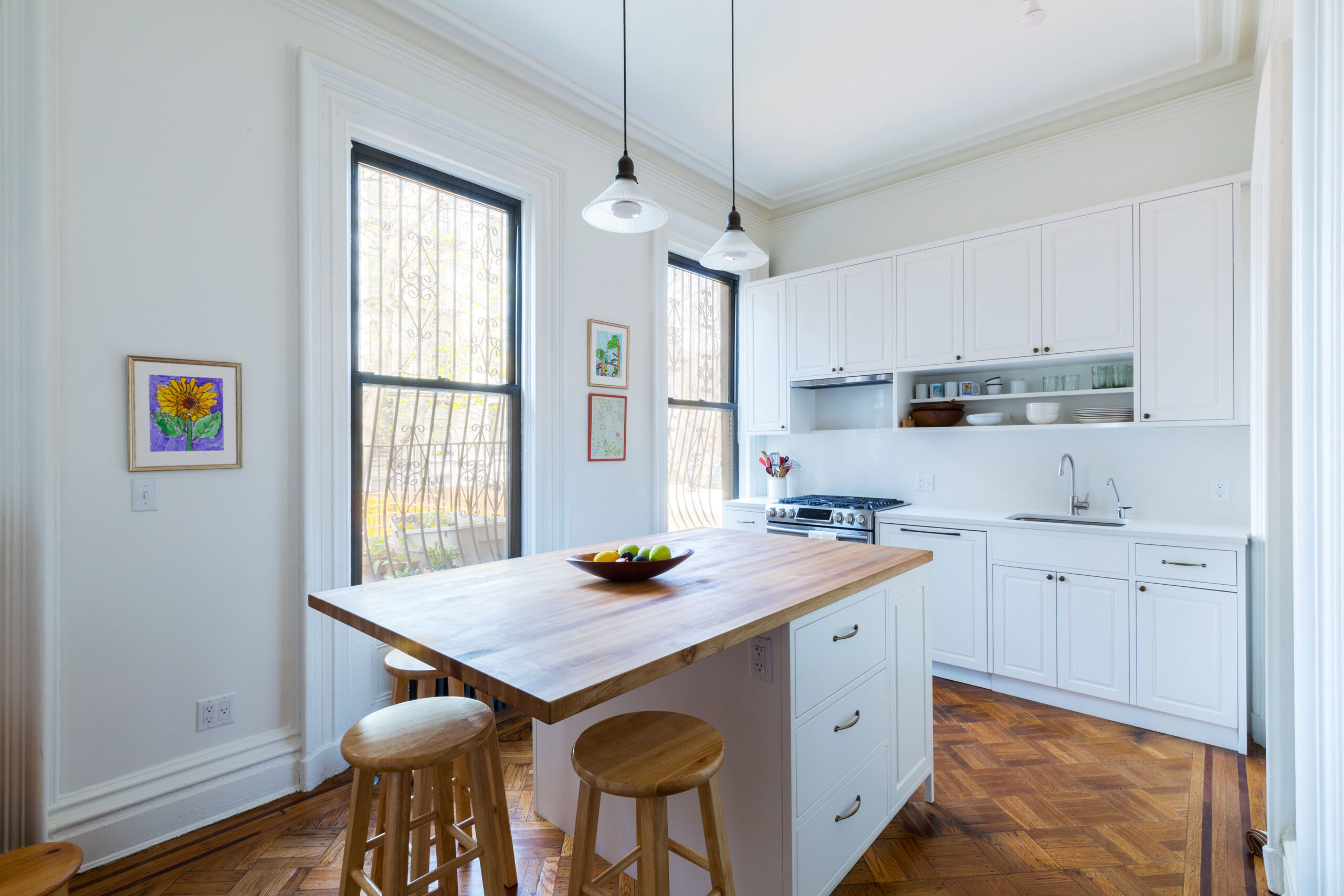 Sunny white kitchen with tall windows, center island topped with butcher block, wood stools and classic patterned parquet flooring
