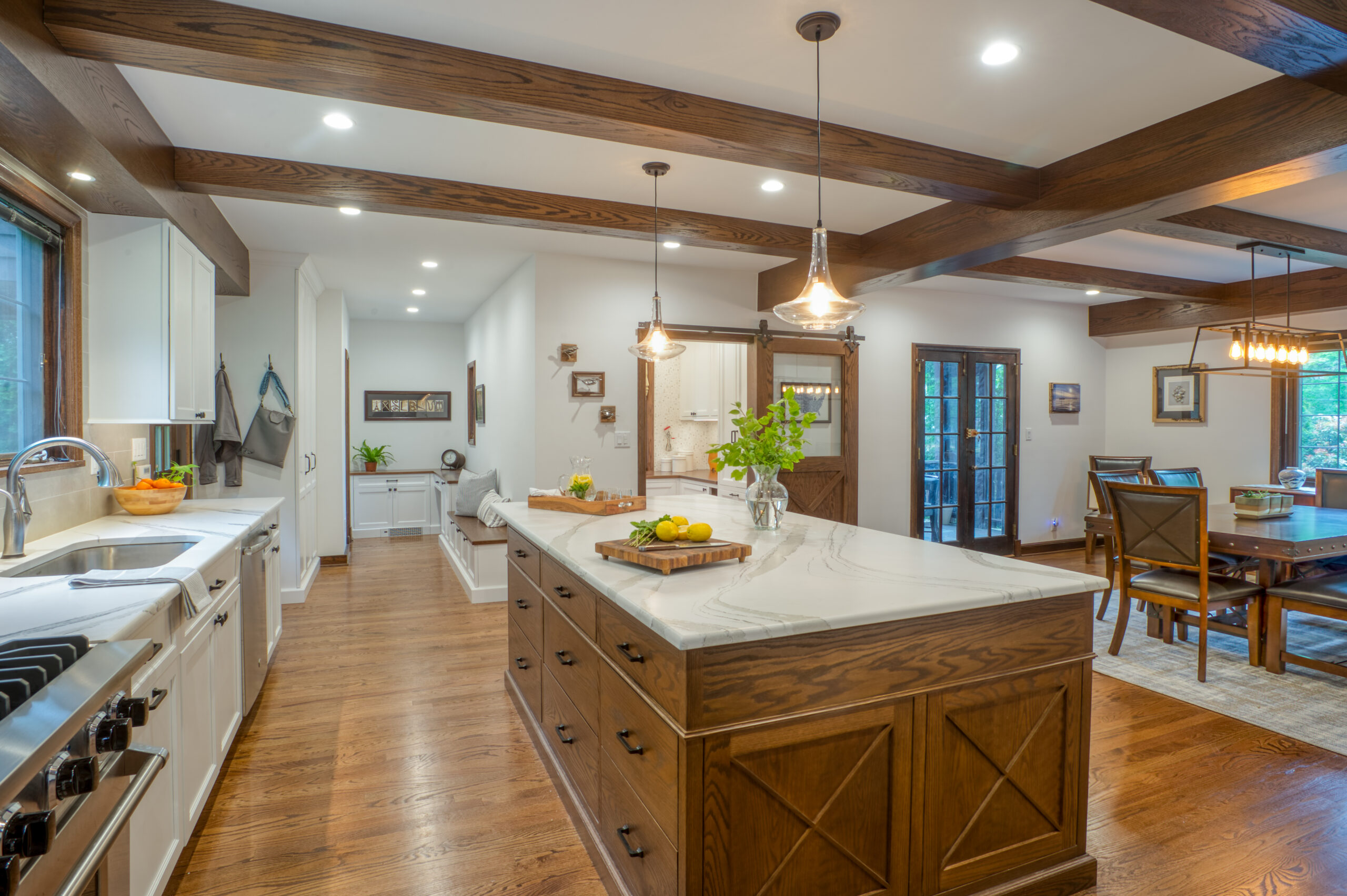Open concept kitchen with a long marble-topped wood island, exposed ceiling beams, and a view into the adjoining dining and sitting areas.