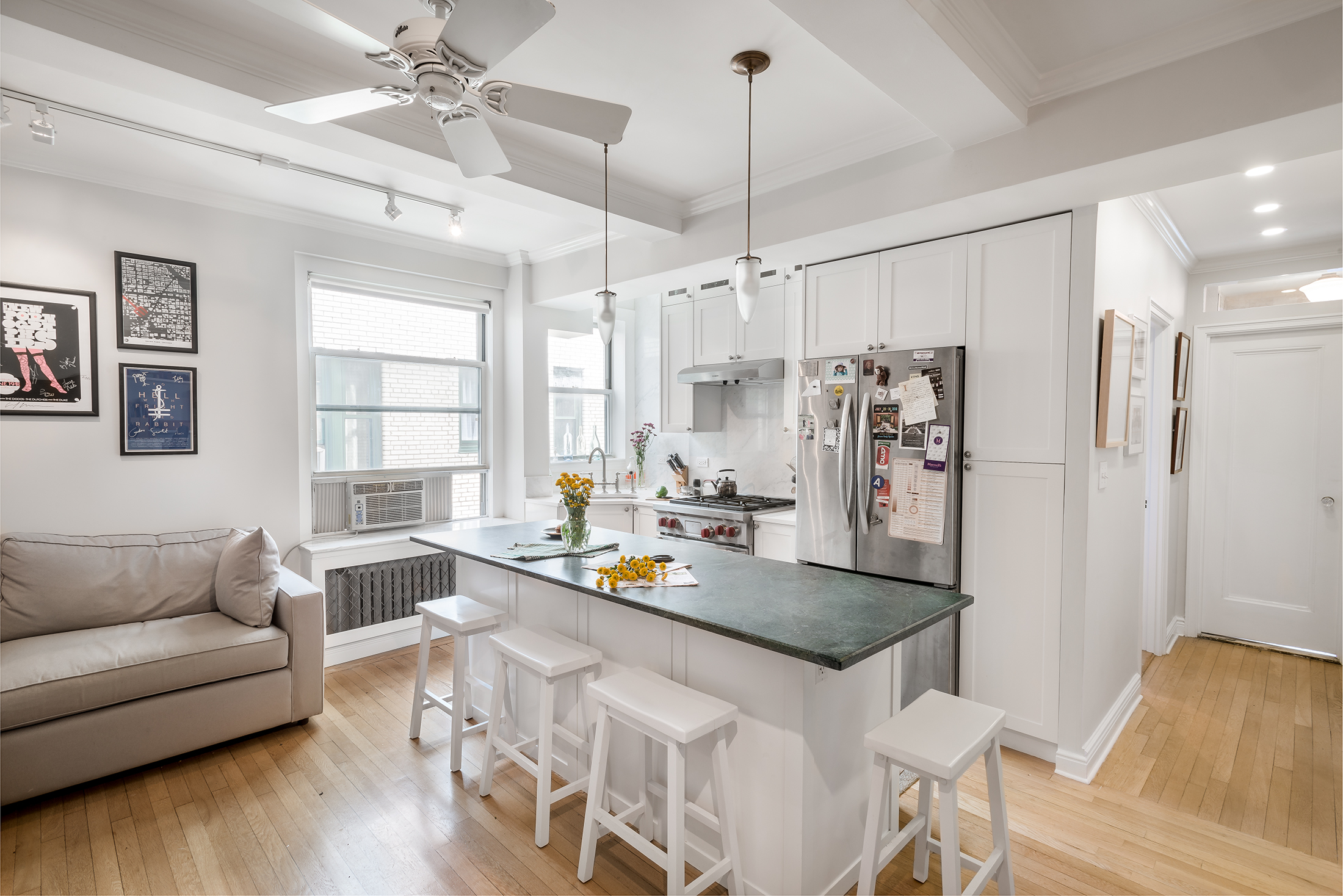 Compact open kitchen with white cabinets, green stone peninsula, white stools, and a sofa nearby under bright windows.