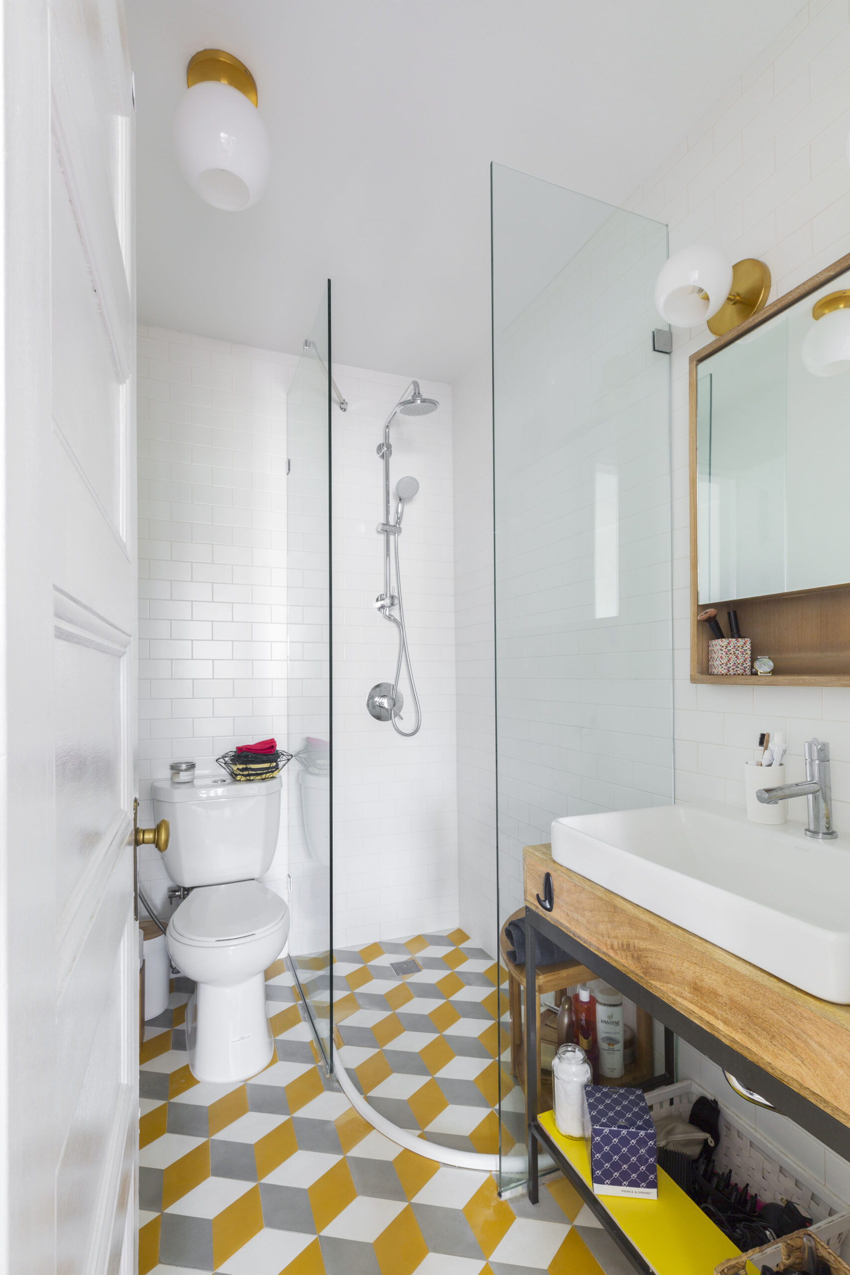 Small bath with frameless glass shower, white subway tile walls, and yellow gray geometric floor beside a wood vanity.