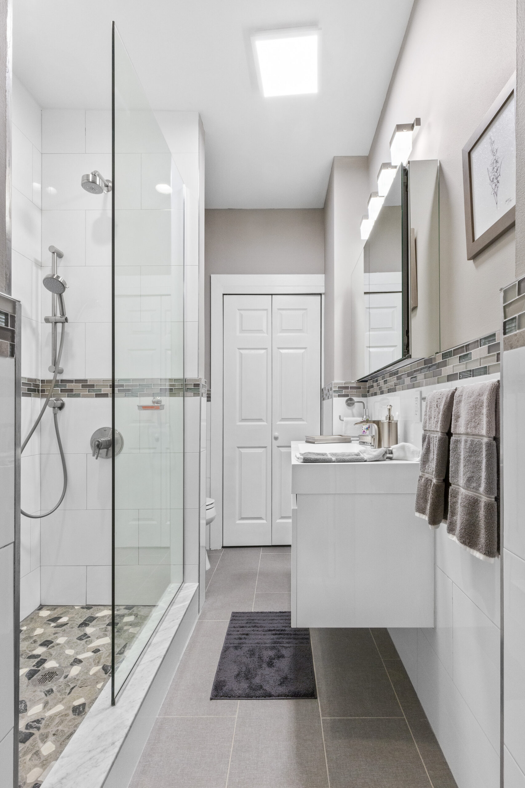Narrow bathroom with glass shower, dual shower heads, large white wall tiles with mosaic band, and a wide white vanity.
