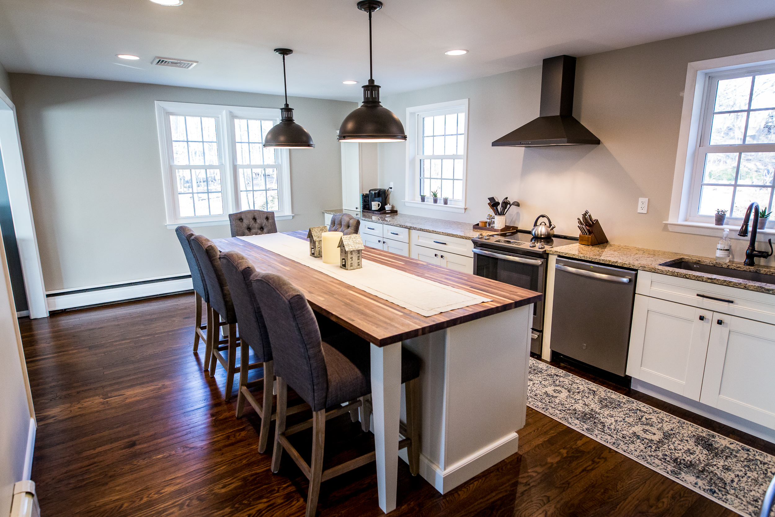 Kitchen with long butcher block island countertop, gray stools, white cabinets, and dark wood floors used for dining and everyday meal prep.