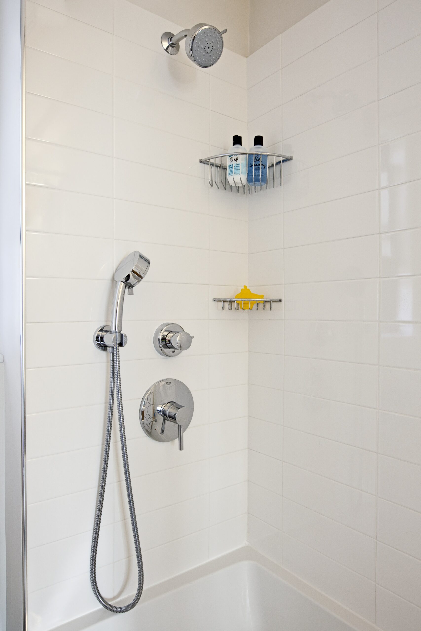 Shower corner with stacked white tile, chrome handheld and wall shower fixtures, corner shelves, and the rim of a drop-in tub.