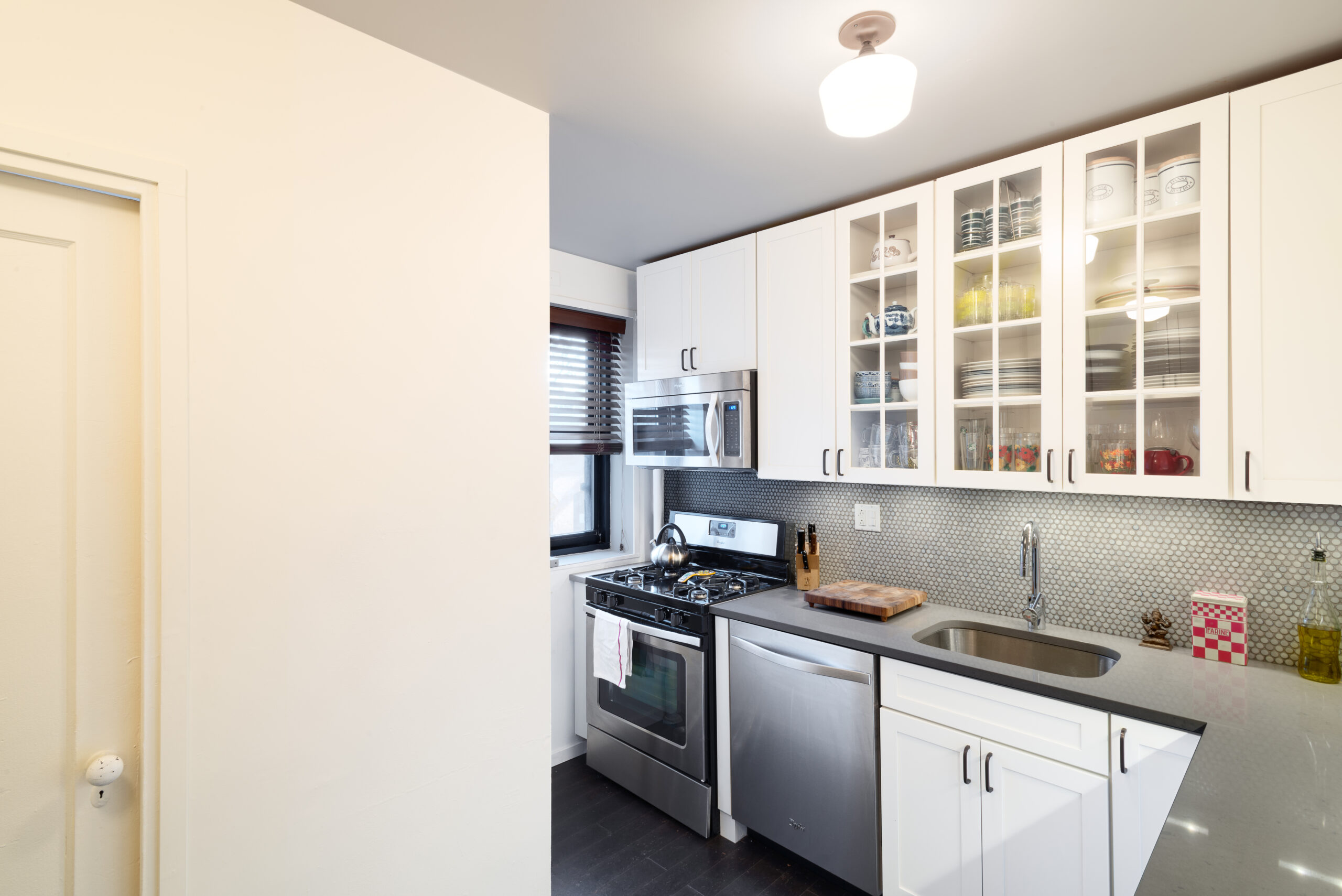 Compact white kitchen with stainless steel stove, microwave, dishwasher, gray counters, glass cabinets, and a wood cutting board adding warmth.