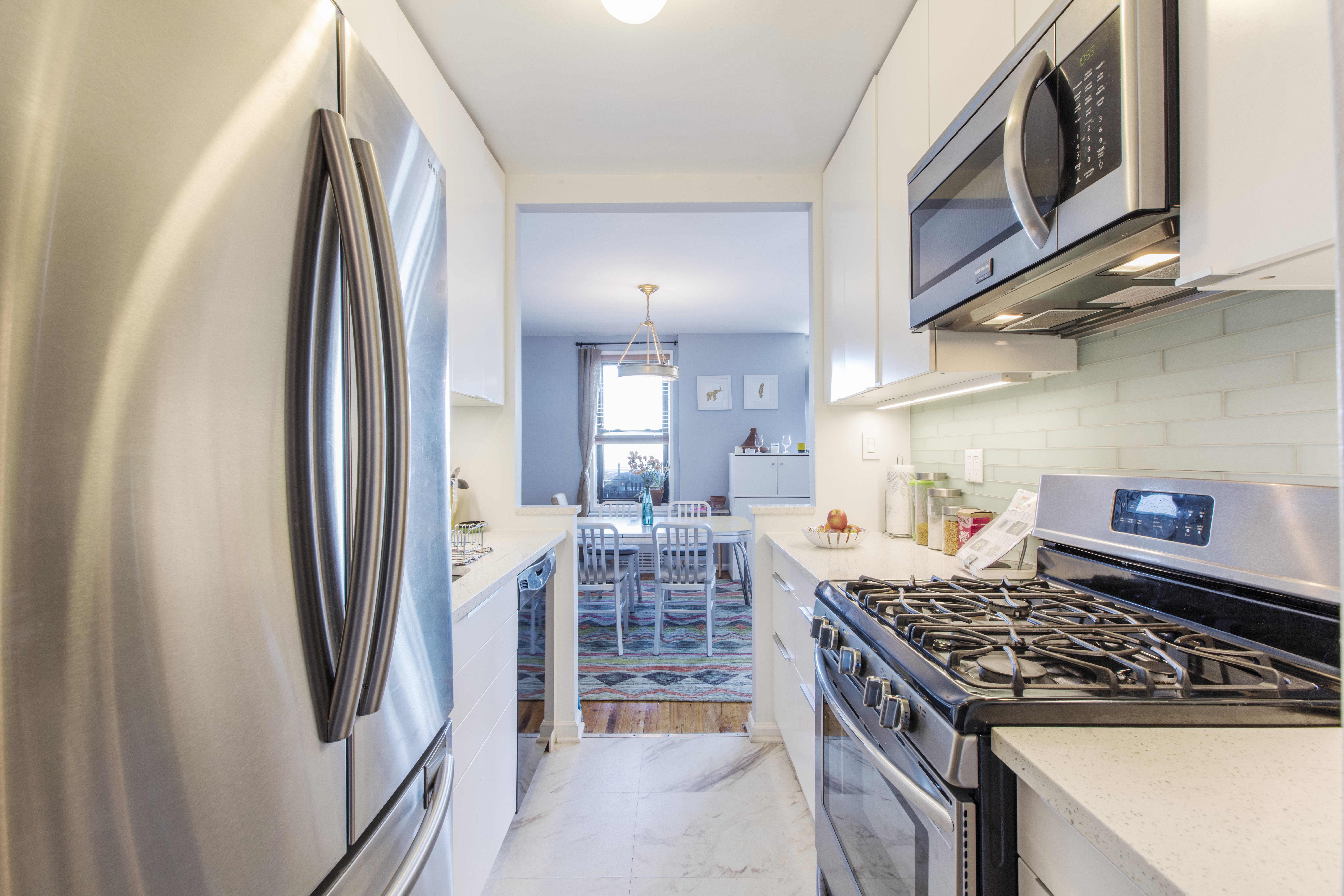 Galley kitchen with stainless steel fridge, range, and microwave, white cabinets, light counters, and view to dining room with warm wood table.
