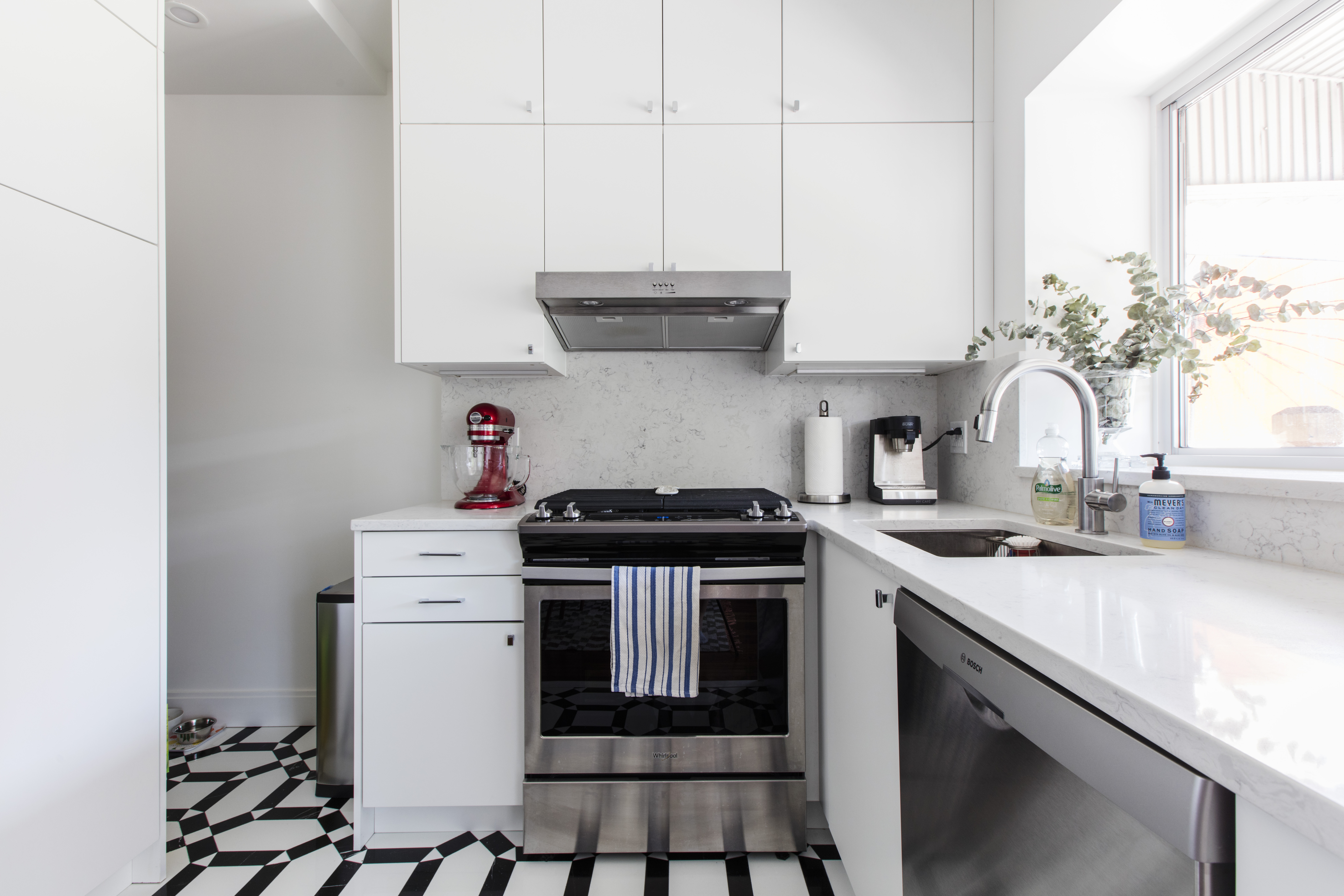 White L shaped kitchen with stainless steel stove, hood, and dishwasher, light counters, and patterned black and white floor tile near a window.
