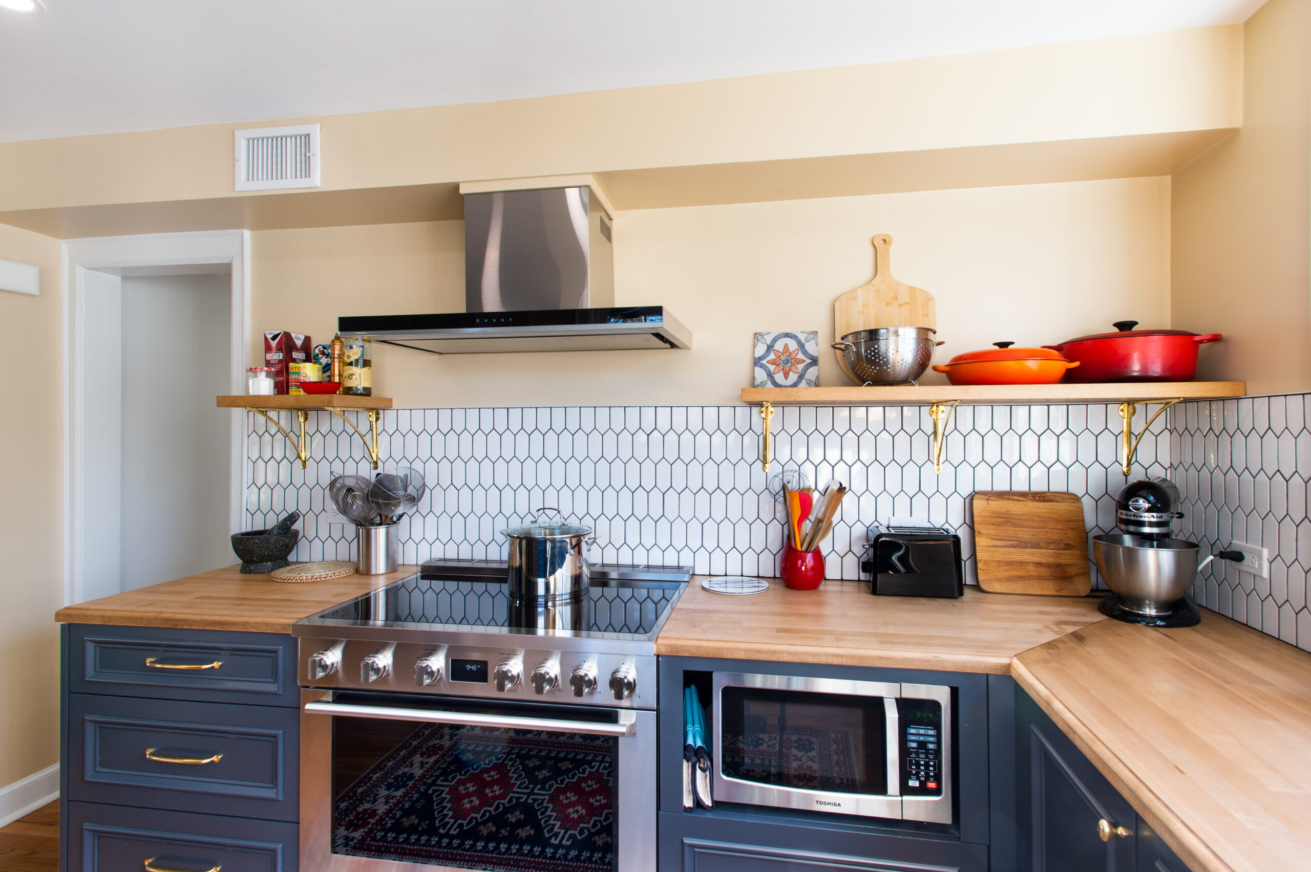 Navy kitchen with stainless steel range and hood, butcher block counters, wood shelves, brass hardware, and geometric tile on the backsplash.