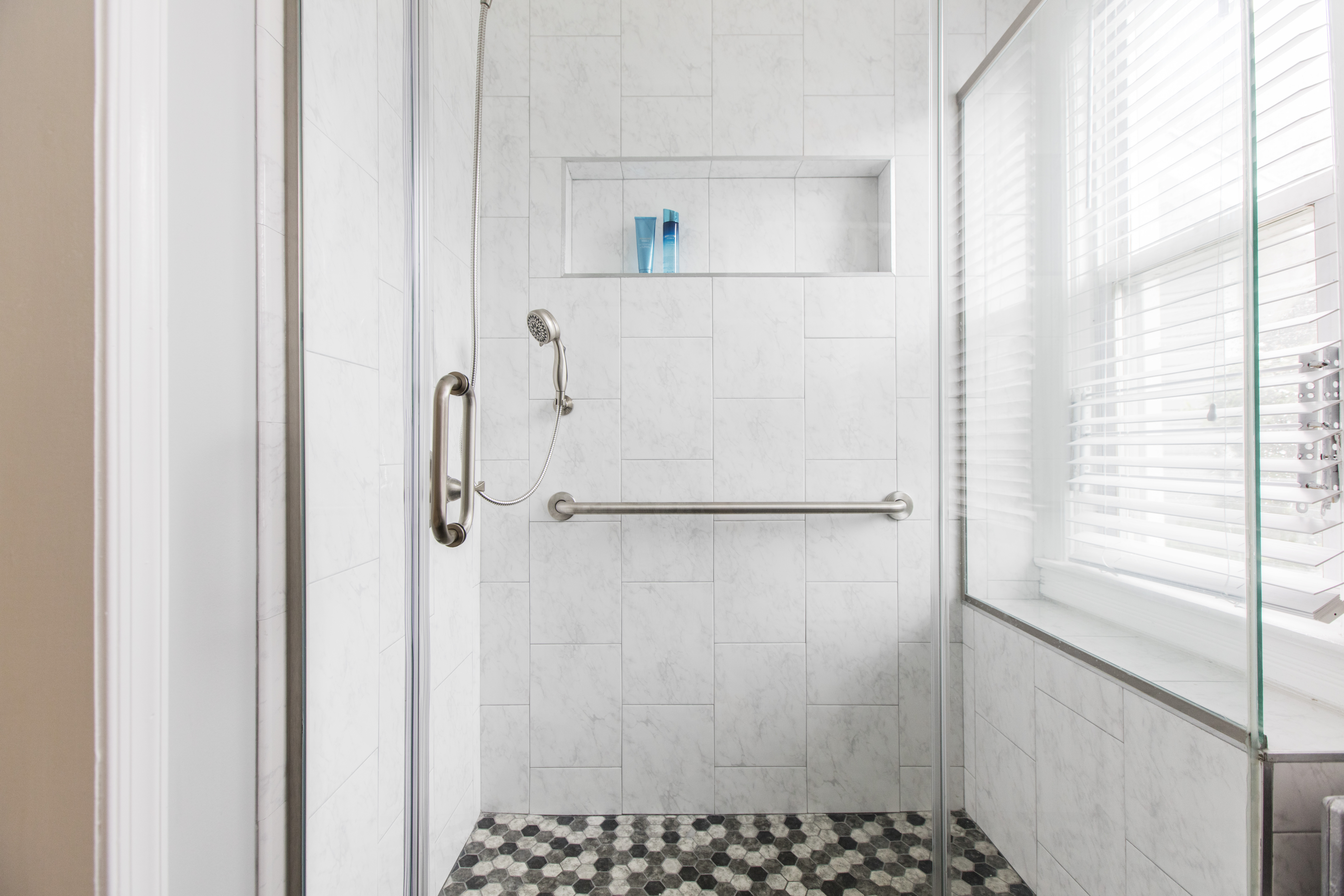 Spacious walk in shower with long grab bar, handheld sprayer, and hex tile floor that provides traction for seniors who need extra stability.