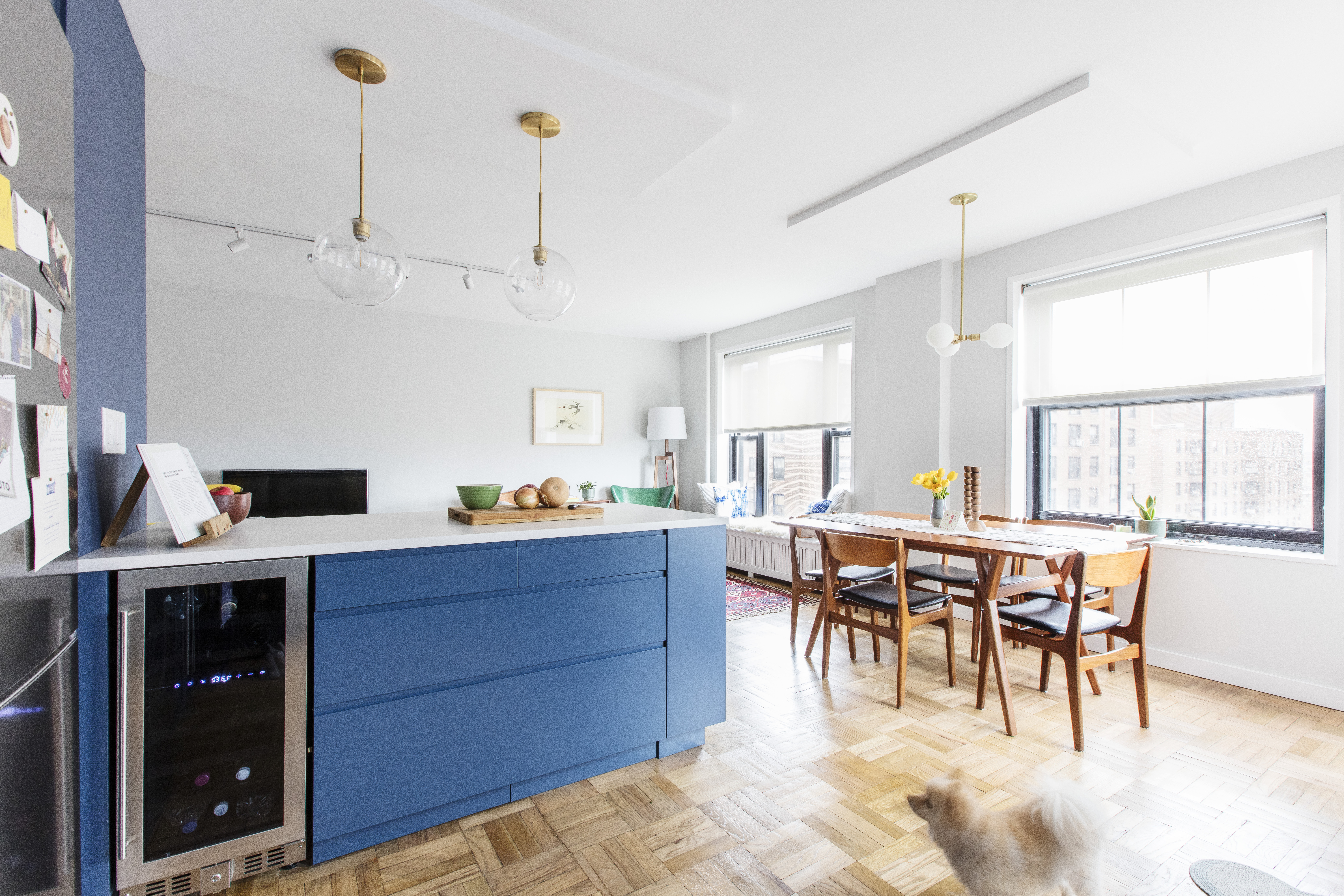 Blue kitchen island with a built-in wine fridge in an open, sunlit apartment dining area with wood flooring.