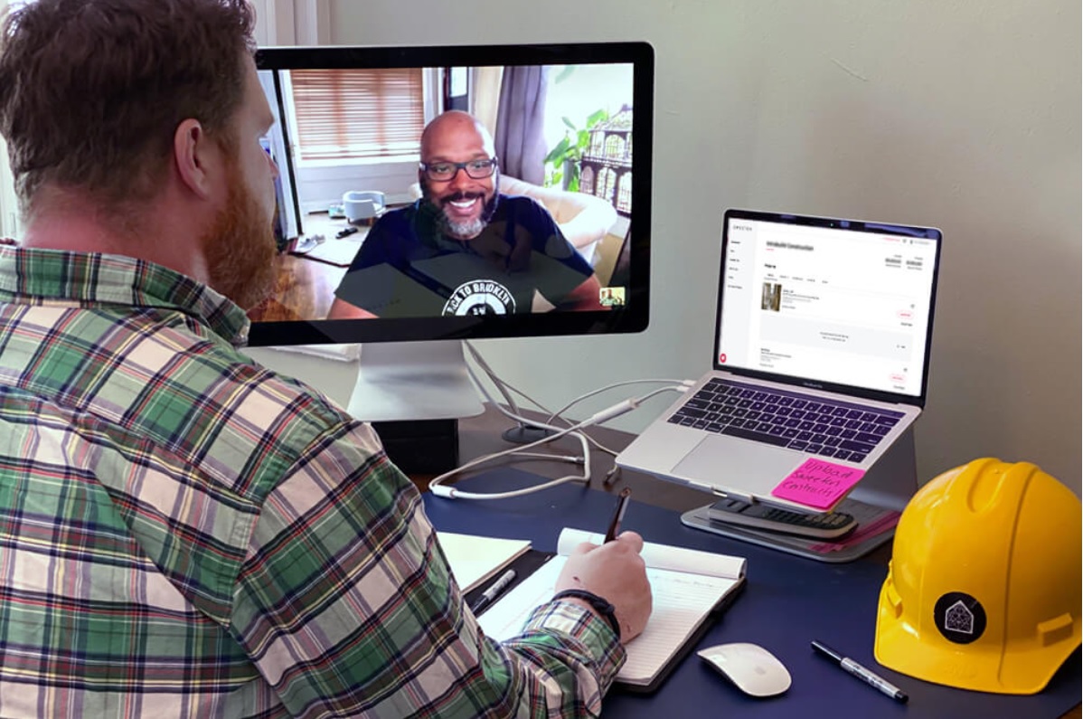 A homeowner meets a contractor through a video call, taking notes beside construction paperwork and a hard hat.