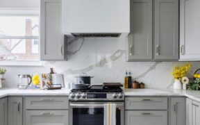 Backsplash slab shows sweeping grey veins, set behind a stainless range, grey shaker cabinetry, brass pulls, and a crisp white hood at center near window.