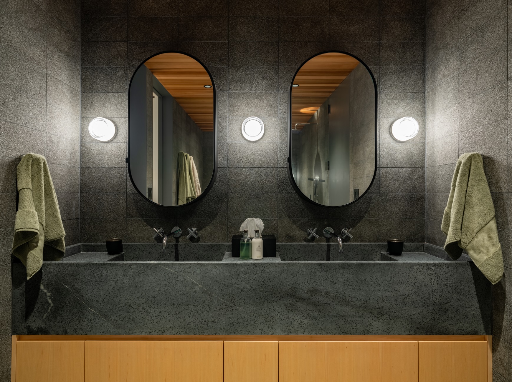 A family’s redesigned bath featuring a maple bathroom vanity, dark stone counter, twin oval mirrors, and wall-mounted faucets.