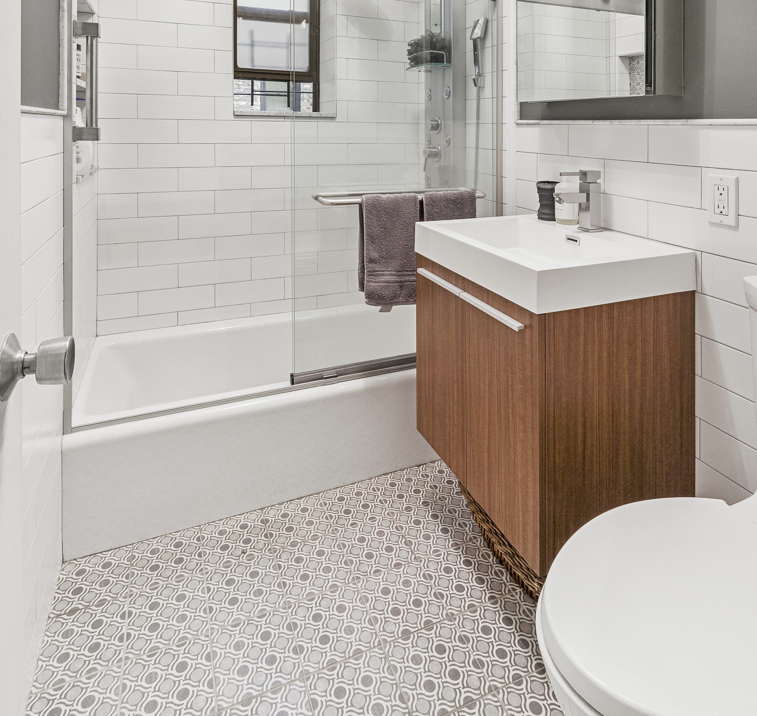 A reworked bath featuring a teak bathroom vanity, white basin, chrome faucet, and patterned floor tile by the tub.