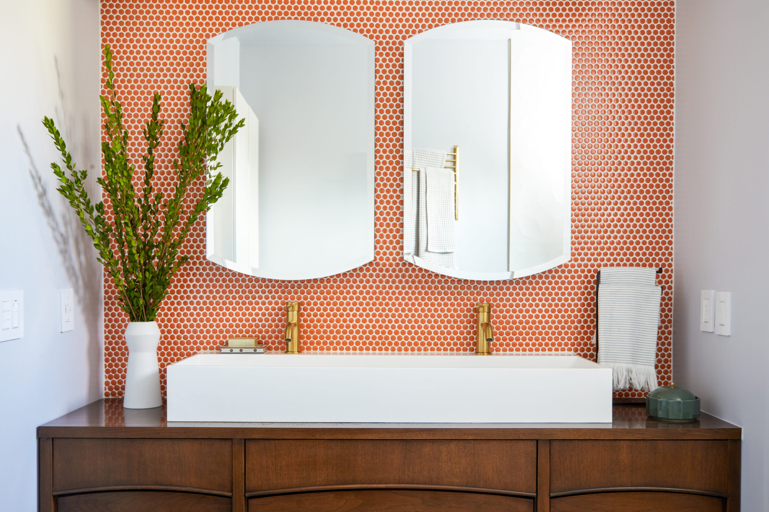 A reimagined bath with a dresser-style bathroom vanity, trough sink, brass faucets, and orange penny-tile wall.