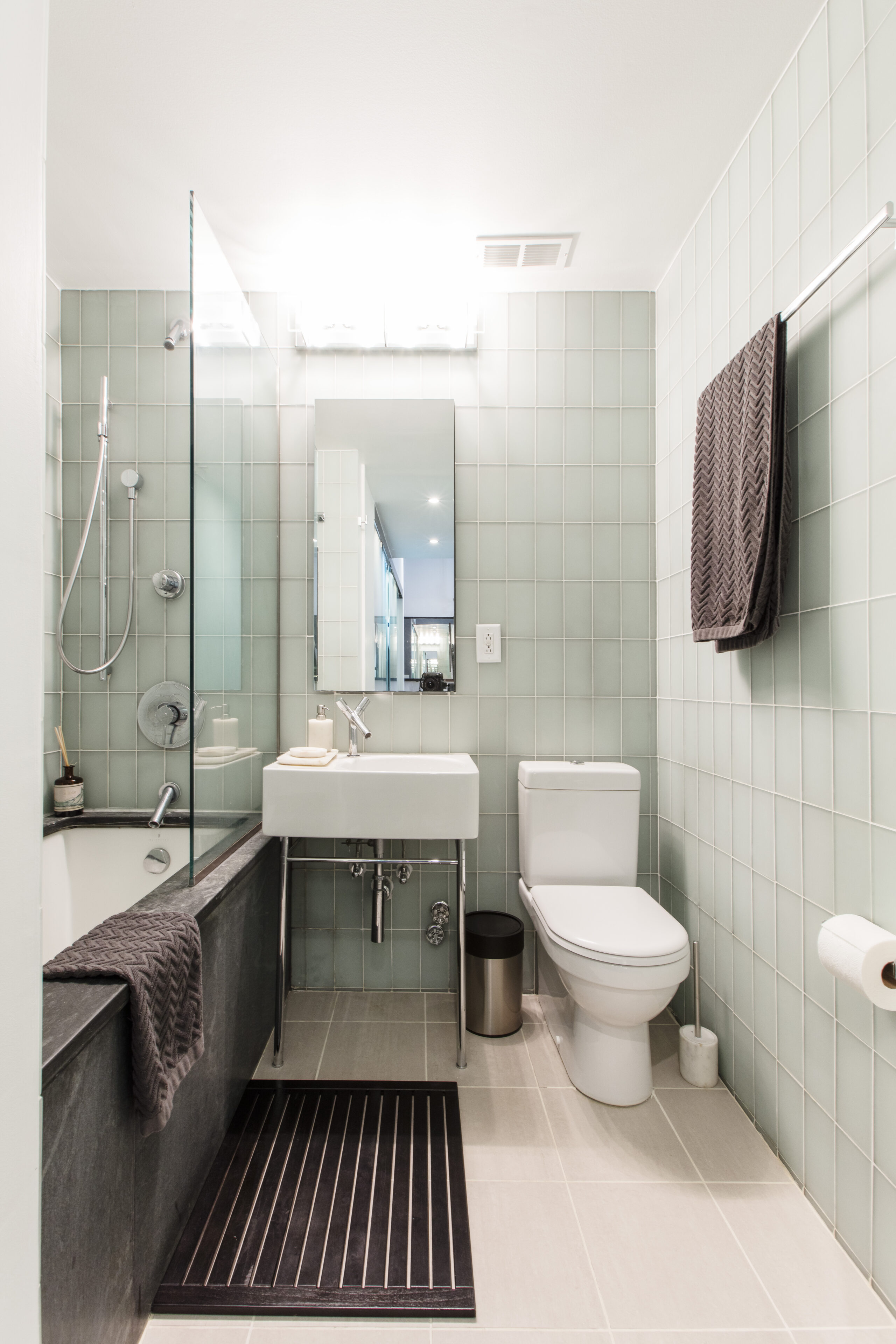 A remodeled bathroom in Manhattan showing light green tile wall, mirror, sink, toilet, and tub with shower