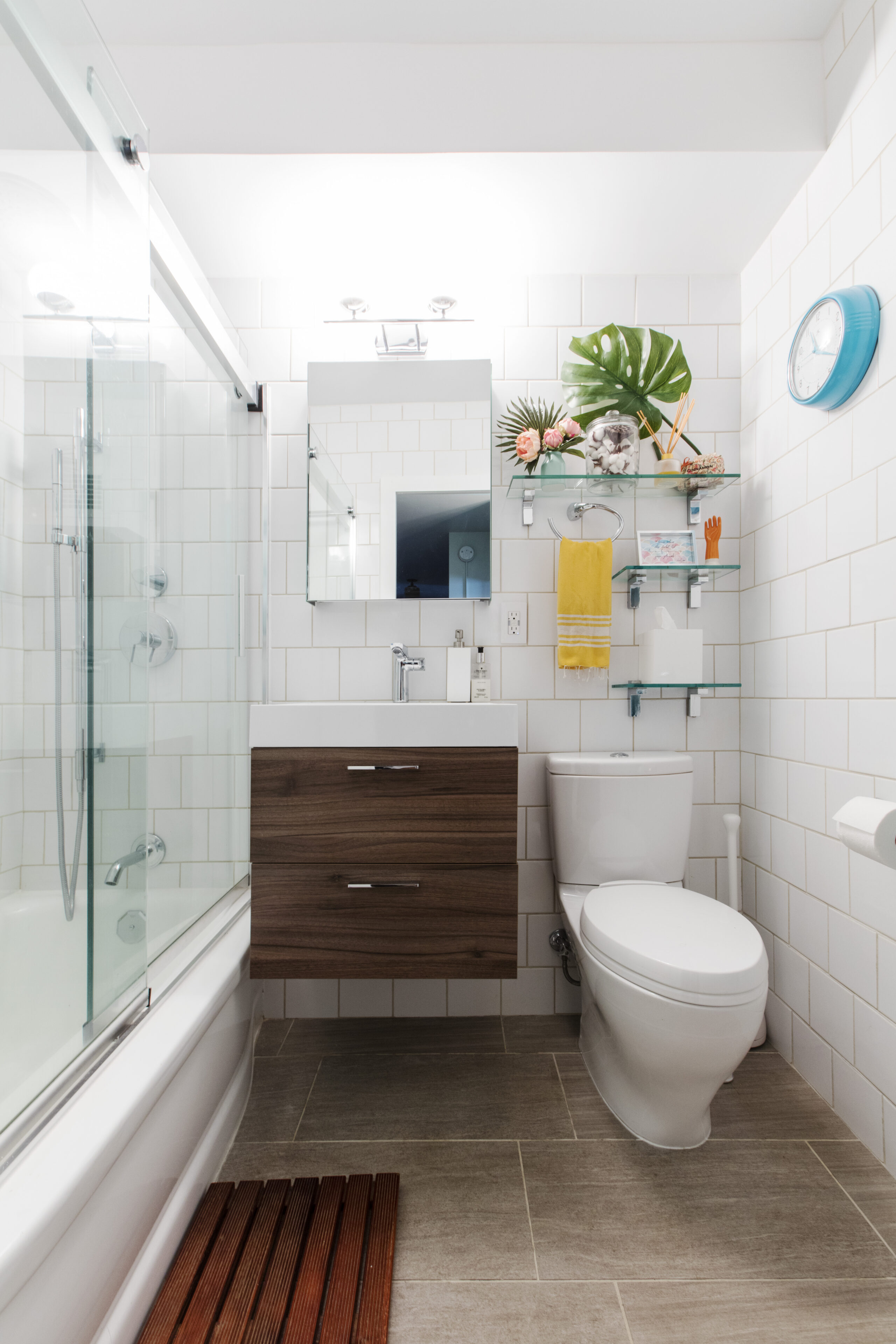 A renovated bathroom in Manhattan with white subway wall tile, floating wood vanity, rectangular mirror, open glass shelves, and glass sliding door in the tub
