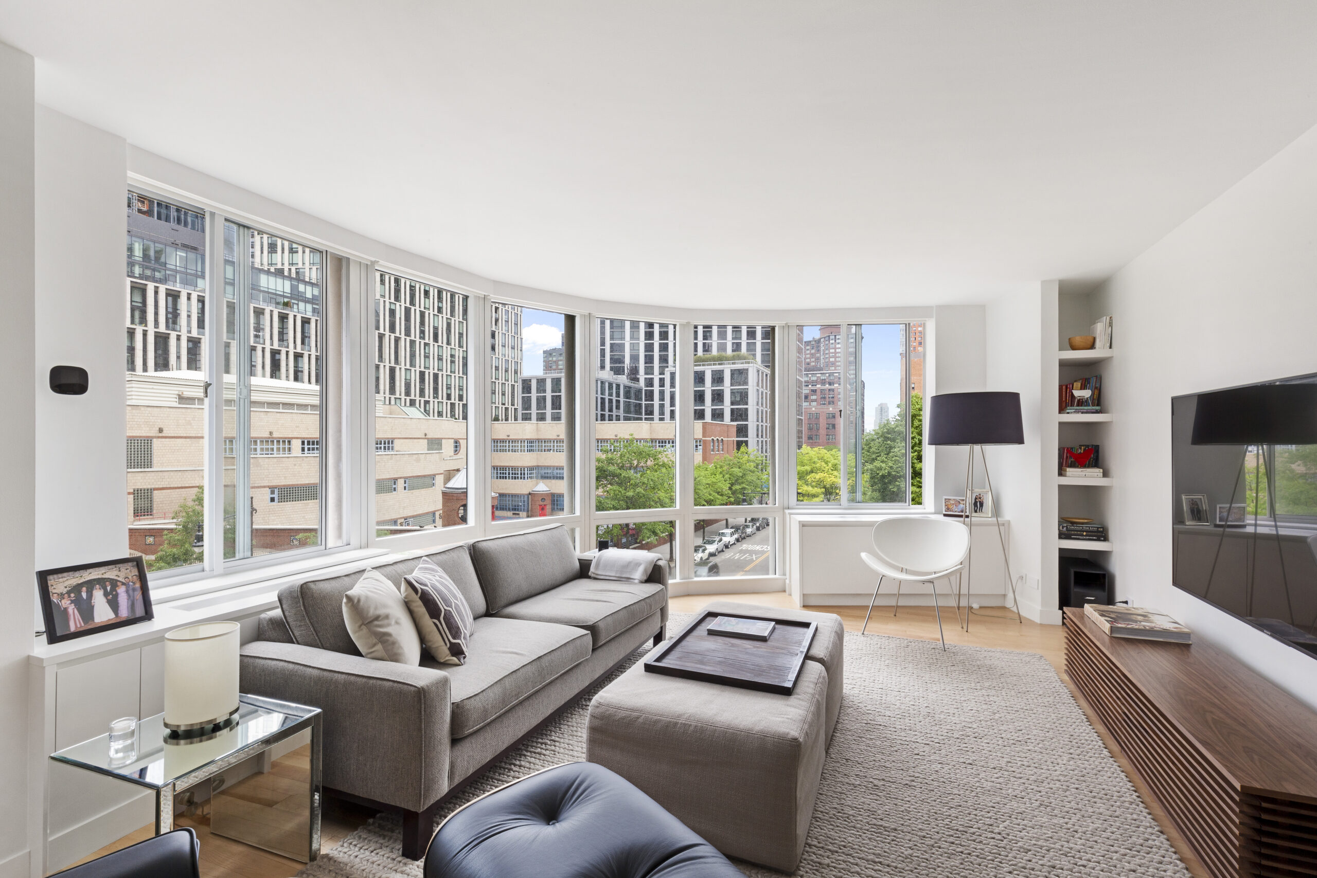 Tribeca living room after renovation with curved windows, wood floors, low media cabinet, built-in shelves, and open flow near the guest room and office combo.