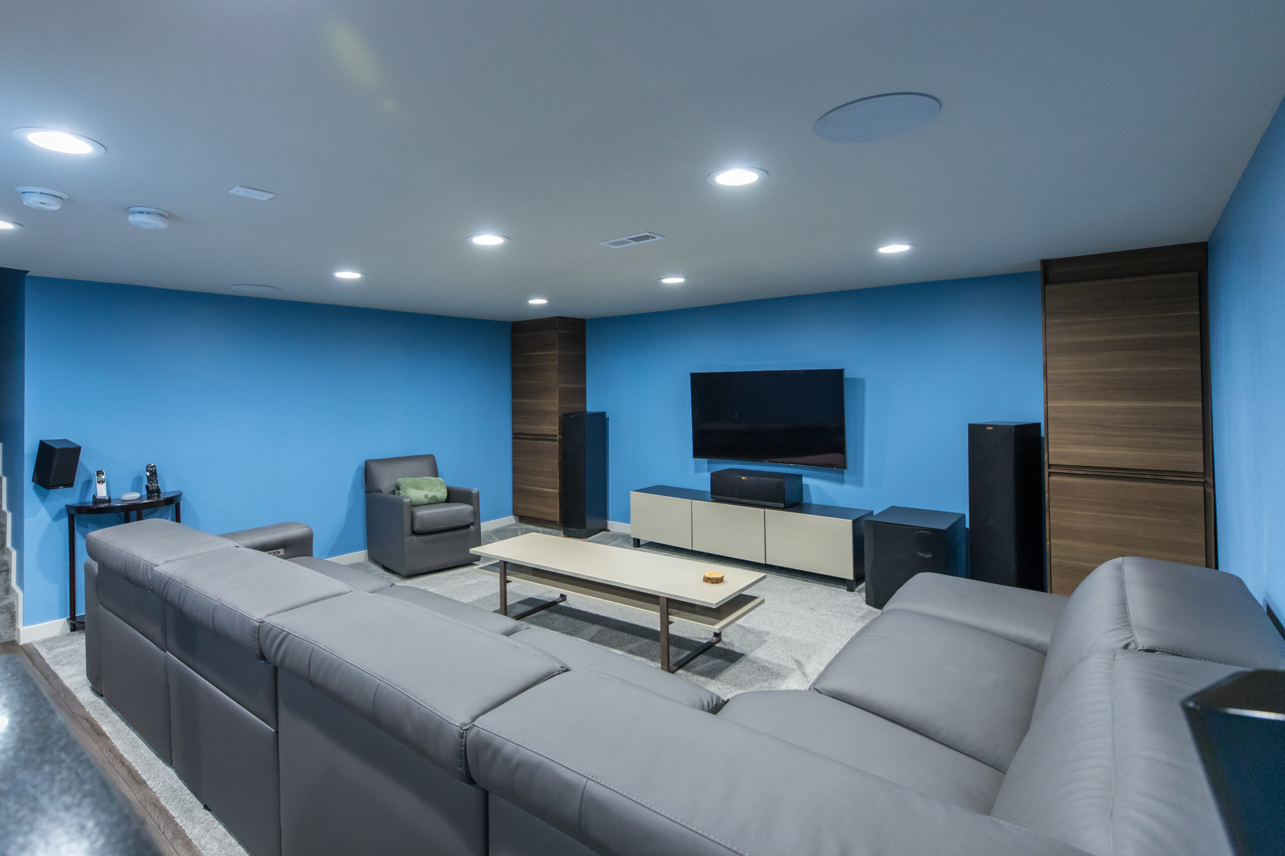 Ardmore basement renovation with blue walls, recessed lighting, gray sectional seating, wall-mounted TV, wood cabinetry, and soft area rug.