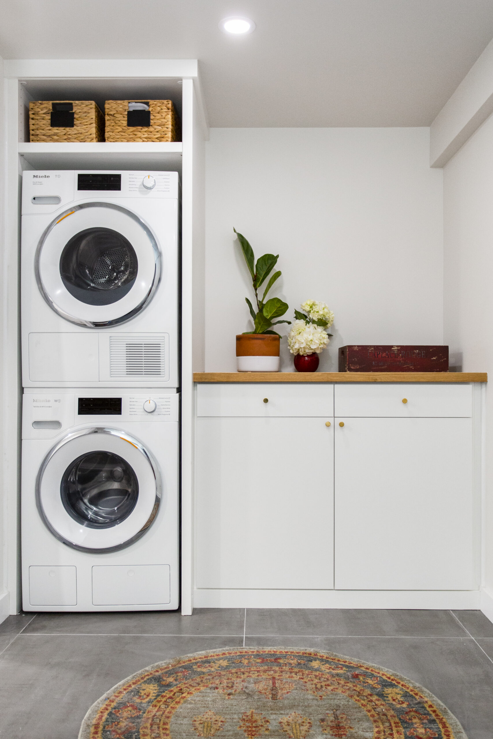 Stuyvesant Heights basement renovation with stacked laundry machines, white cabinets, wood countertop, storage baskets, gray tile, and recessed light.