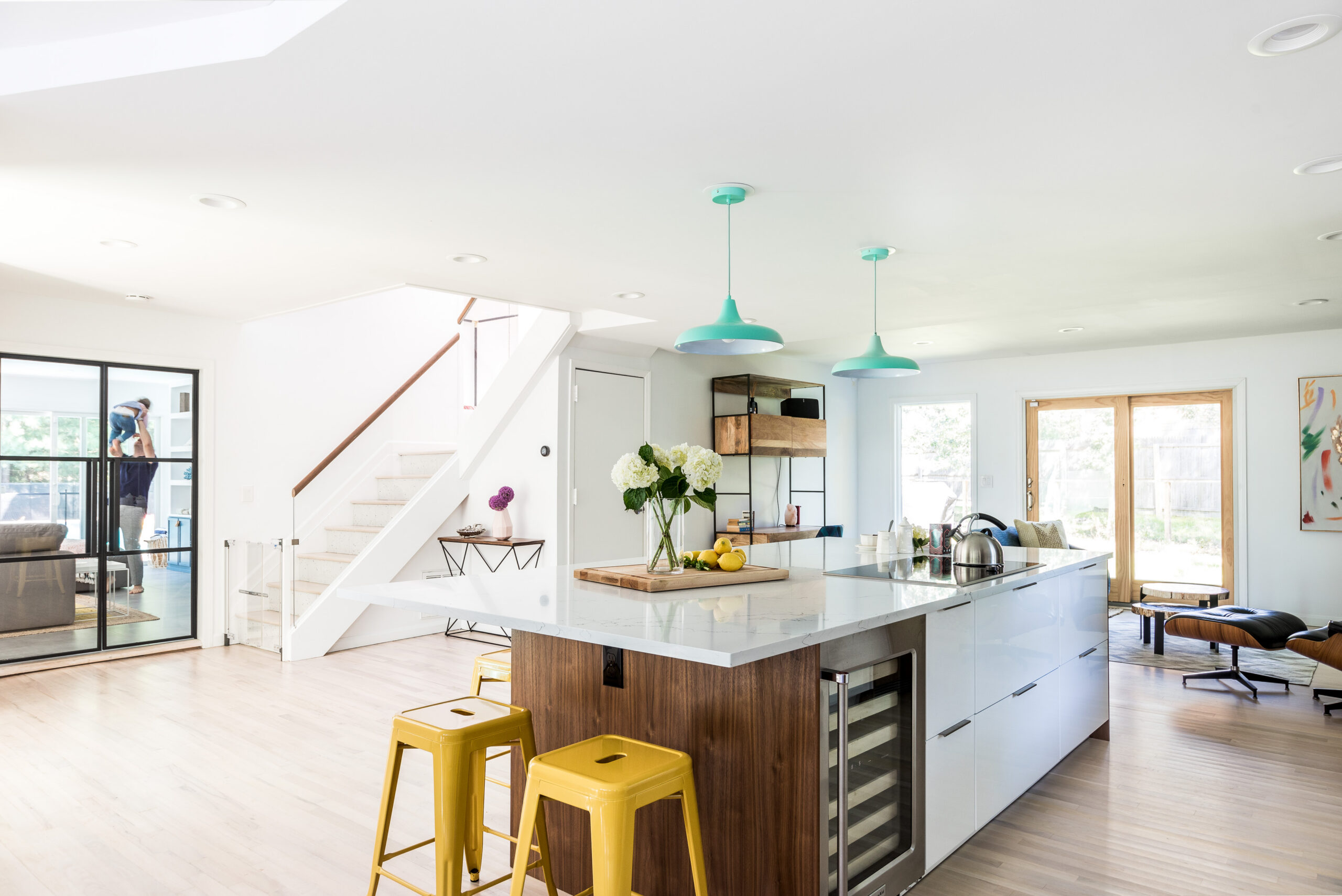 East Hampton open kitchen with large quartz island, walnut panels, glossy white cabinets, aqua pendants, yellow stools, and connected living areas.
