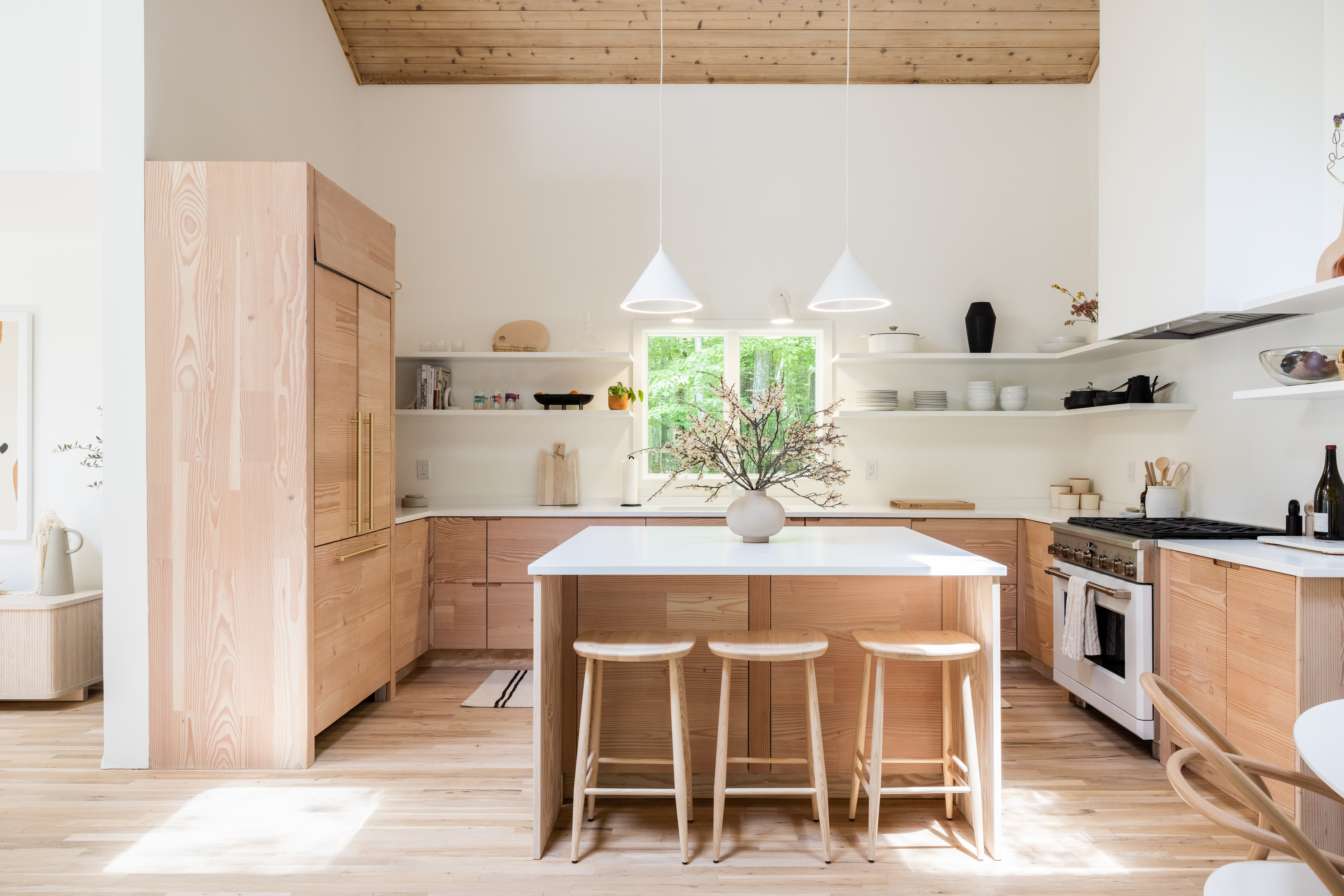 Amagansett beach house kitchen with Douglas fir cabinets, white island, open shelving, wood ceiling, pendant lights, and pale floors.