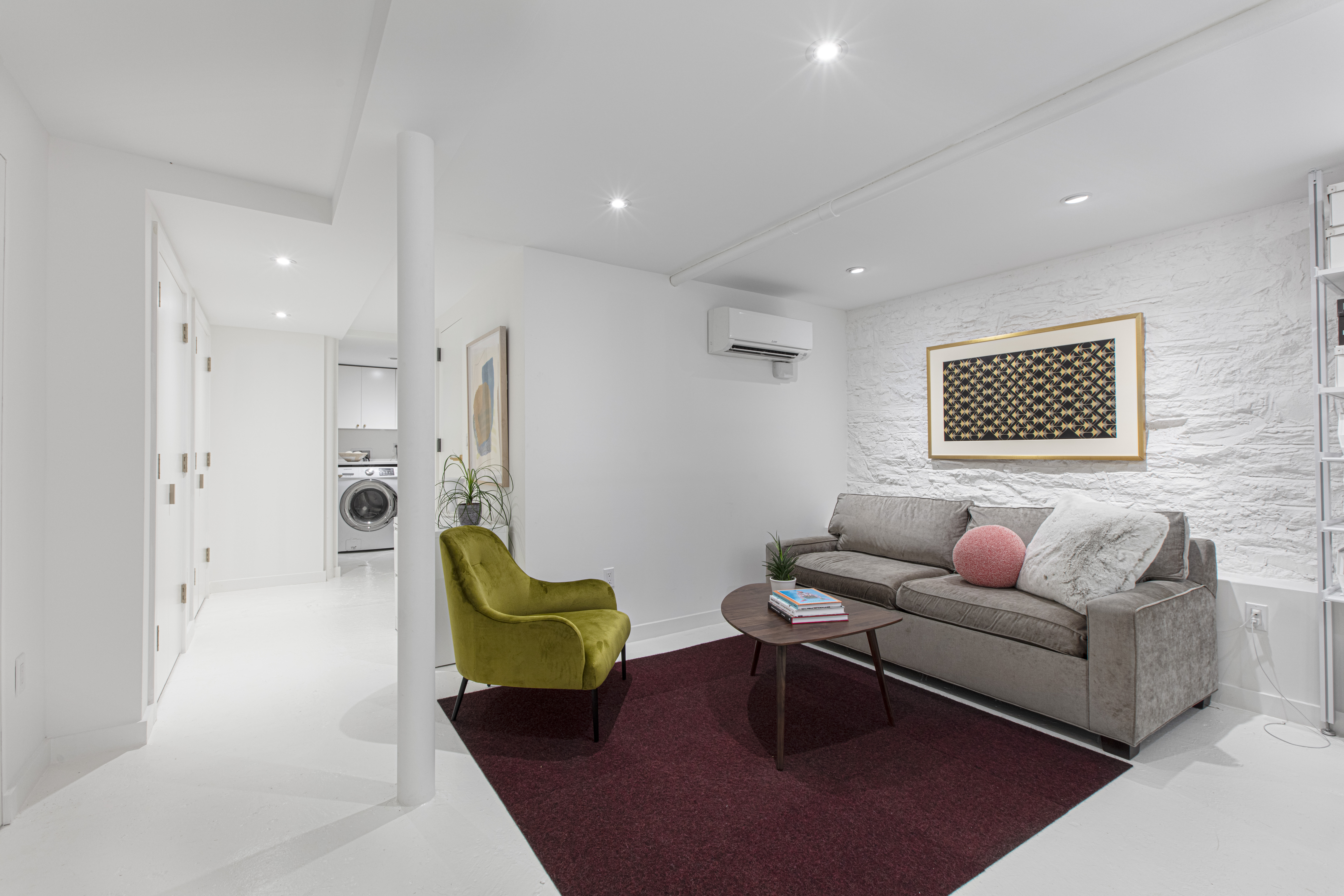 Stuyvesant Heights basement renovation with painted brick, recessed lighting, gray sofa, green lounge chair, red rug, and laundry area beyond.