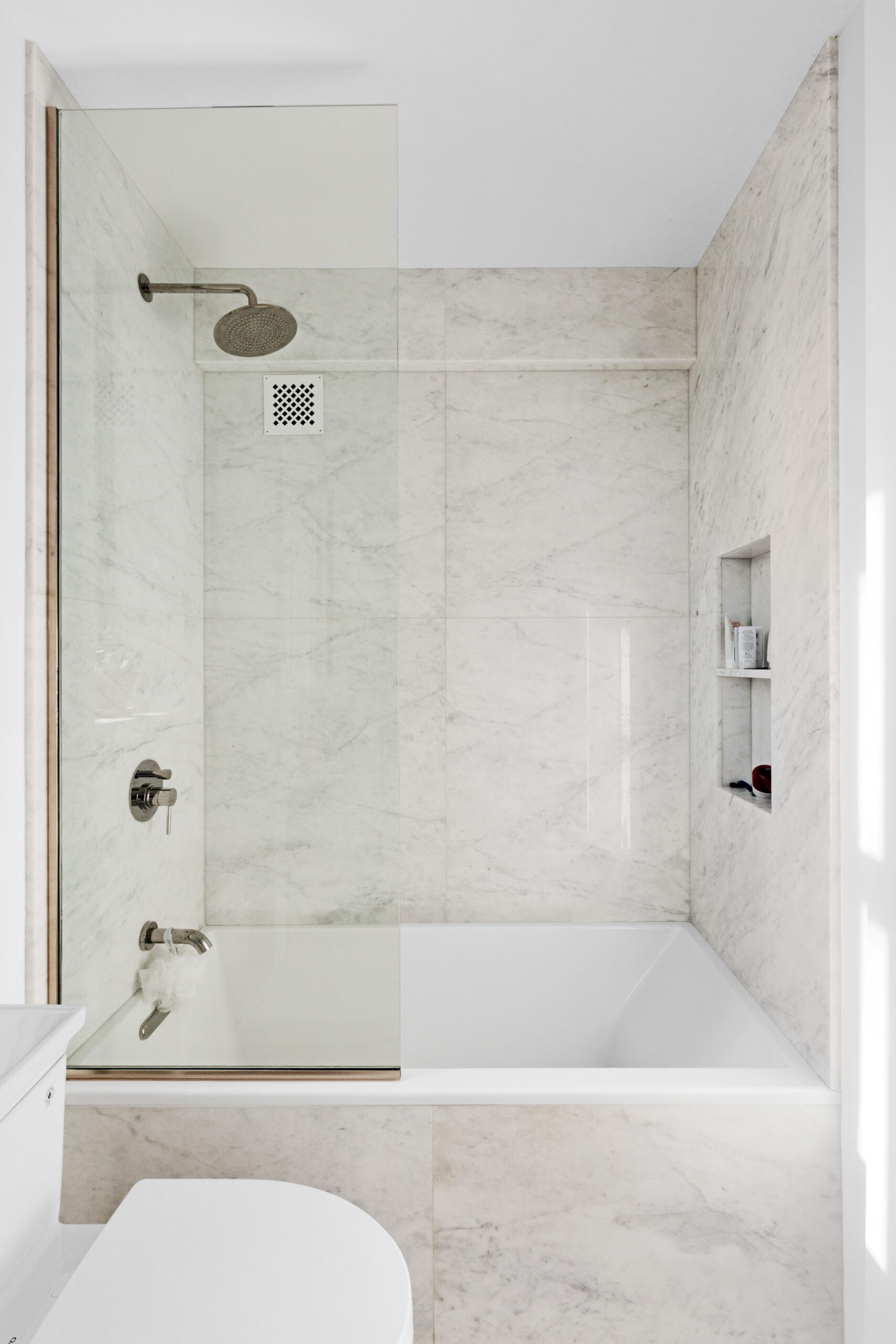 Chelsea Marble Bathroom with marble tub surround, glass panel, deep tub, rain showerhead, and built-in niche for streamlined bath storage.