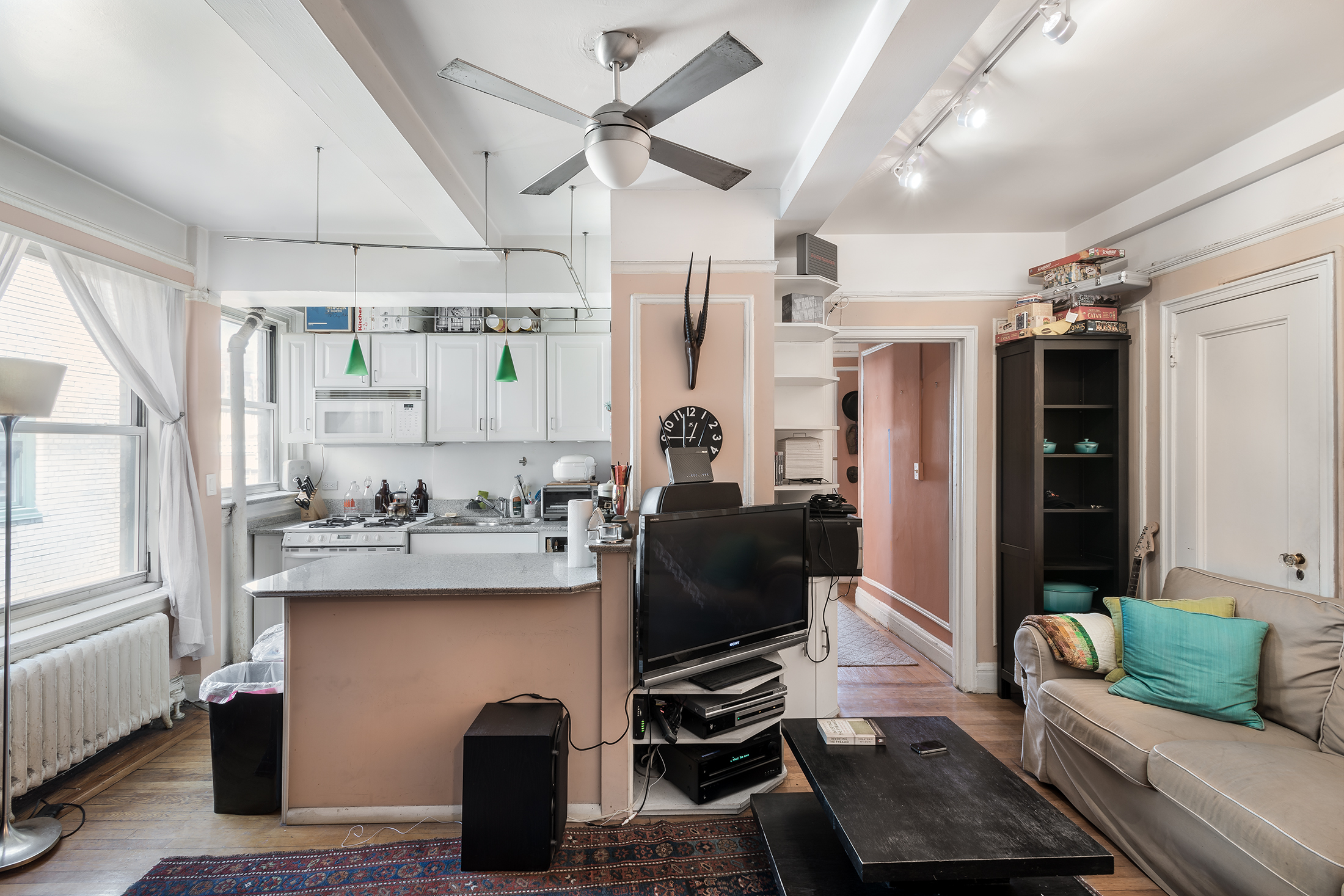 Hell’s Kitchen apartment before renovation with a compact kitchen pass-through, partial wall, track lighting, and living area divided from the workspace.