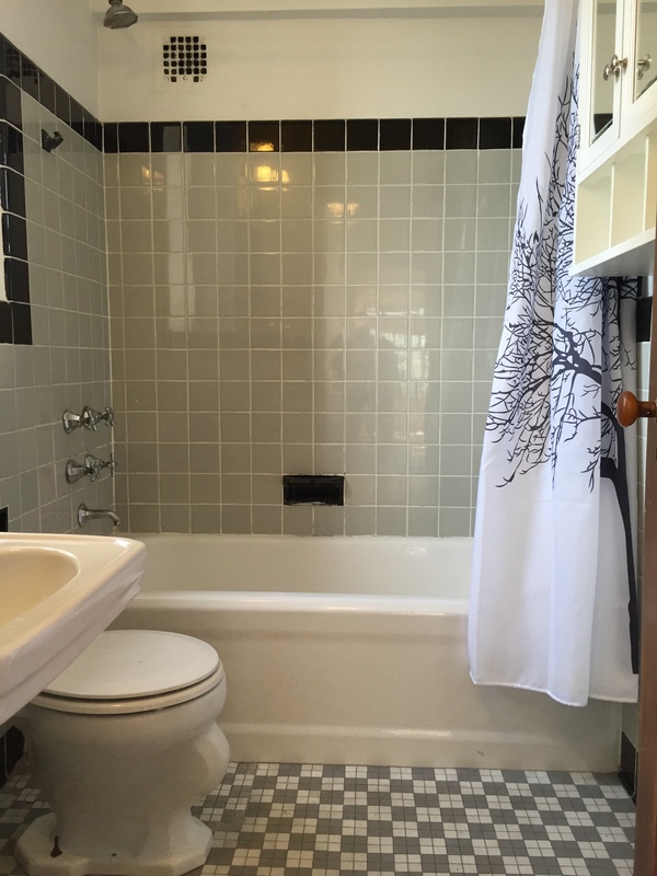New York City bathroom before renovation with dated wall tile, dark border tile, mosaic floor, exposed fixtures, and a shower curtain.