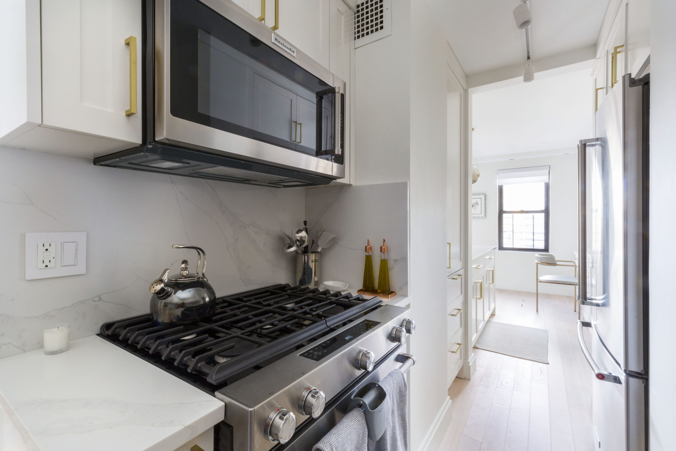 Renovated Upper East Side kitchen with white cabinets, brass pulls, marble-style backsplash, stainless range, and a view toward the kitchen desk niche.