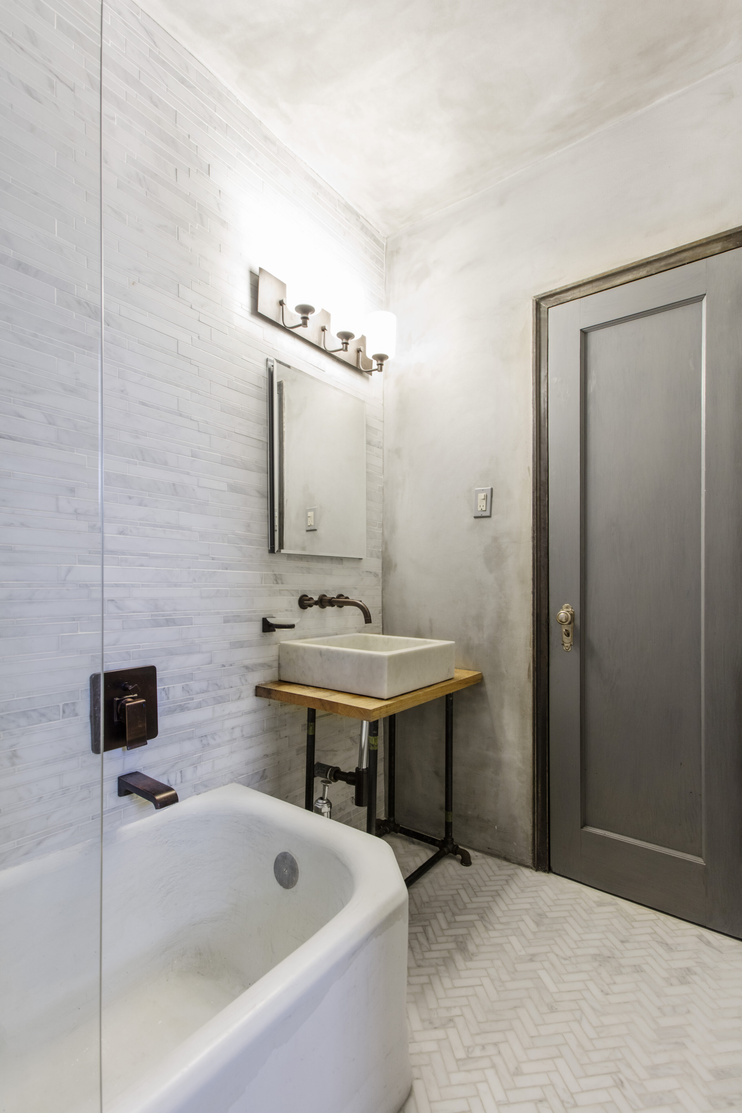 A Brooklyn wabi-sabi inspired bathroom design showing a Carrara marble sink and walls, herringbone floor tile, and white tub.