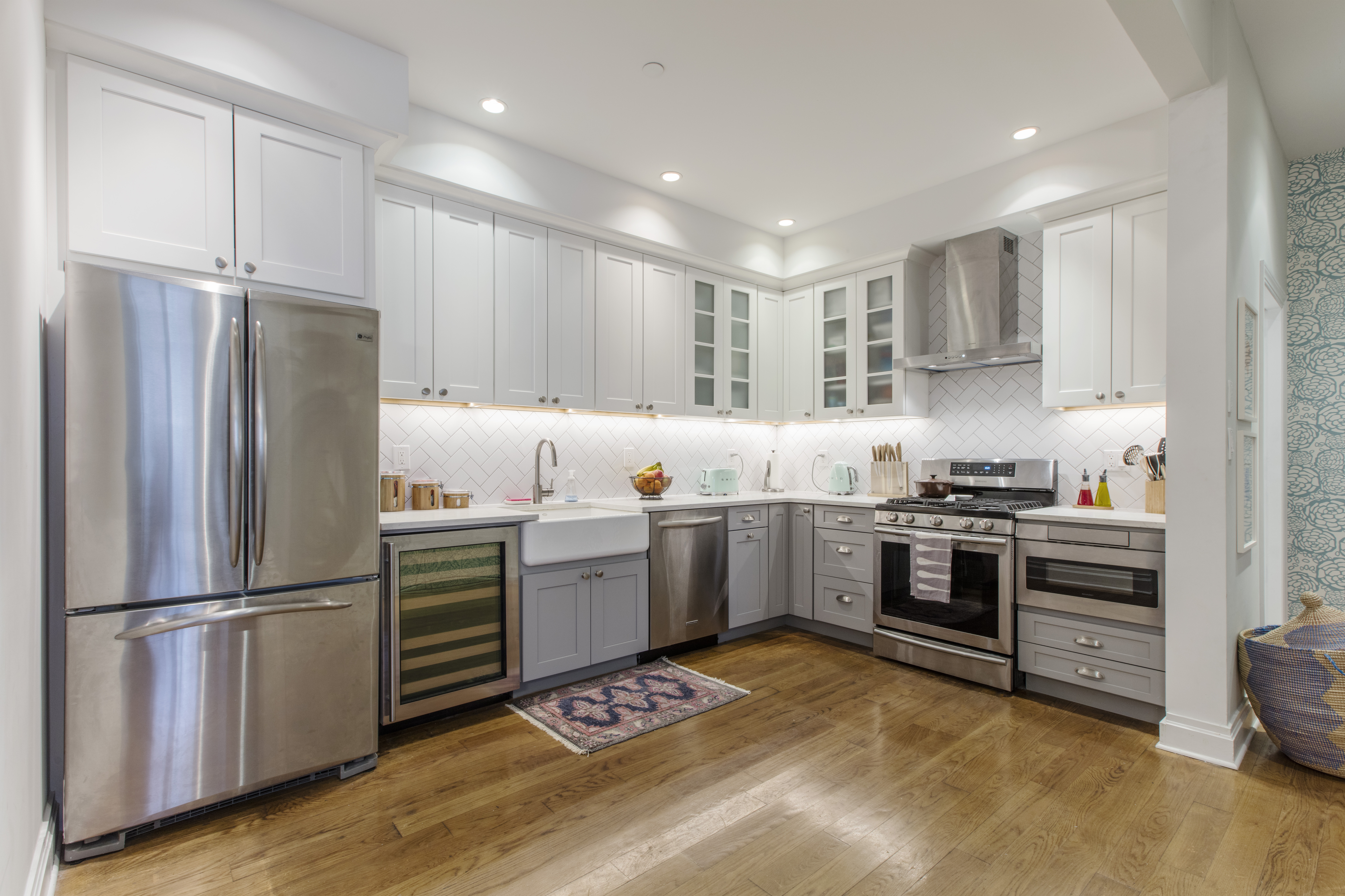 Carroll Gardens kitchen with white and gray shaker cabinets, herringbone tile, wine fridge, farmhouse sink, range hood, and warm wood floors.