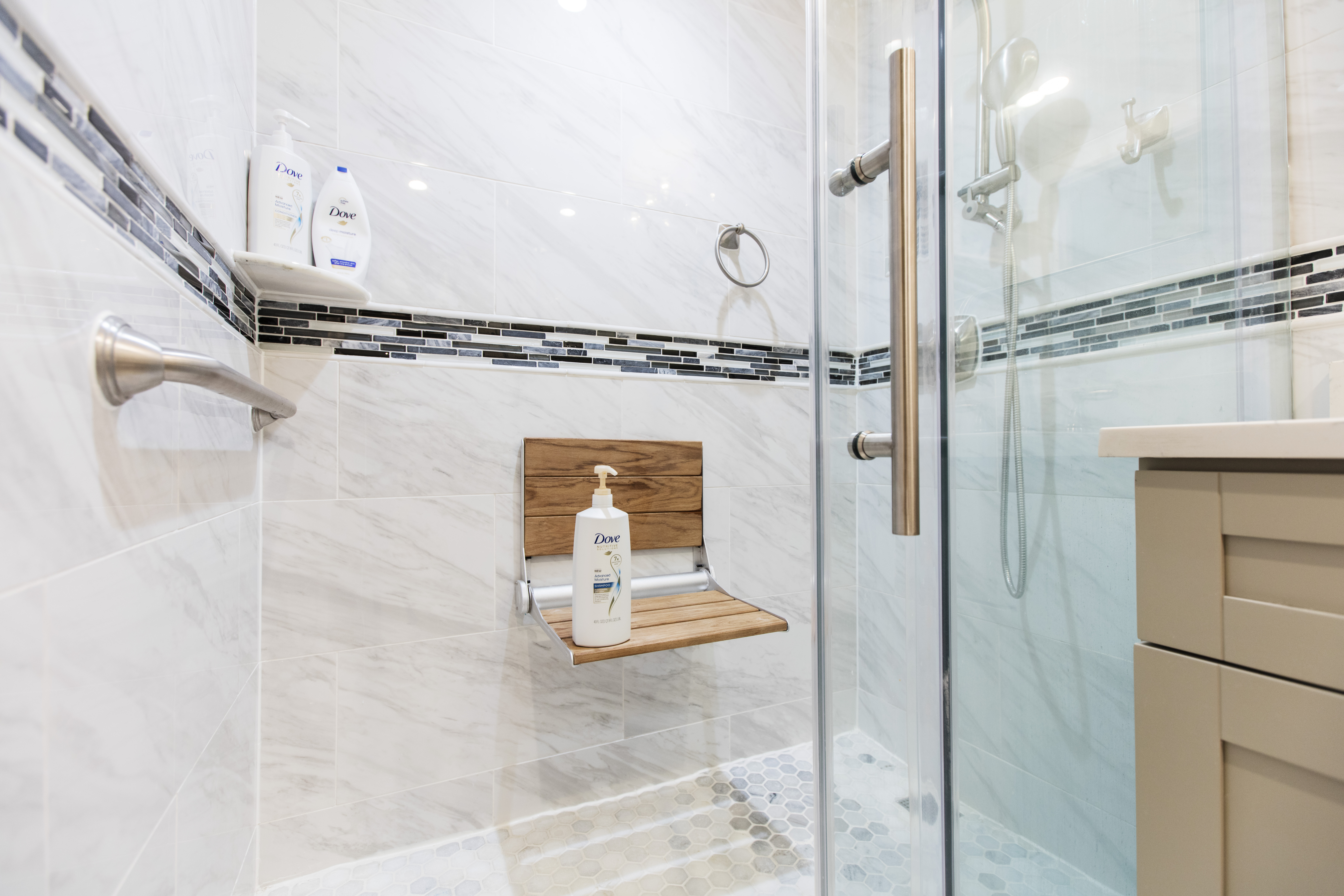 Bronx shower with a fold-down wood seat, grab bar, handheld shower, glass door, marble-look wall tile, and hex mosaic flooring.